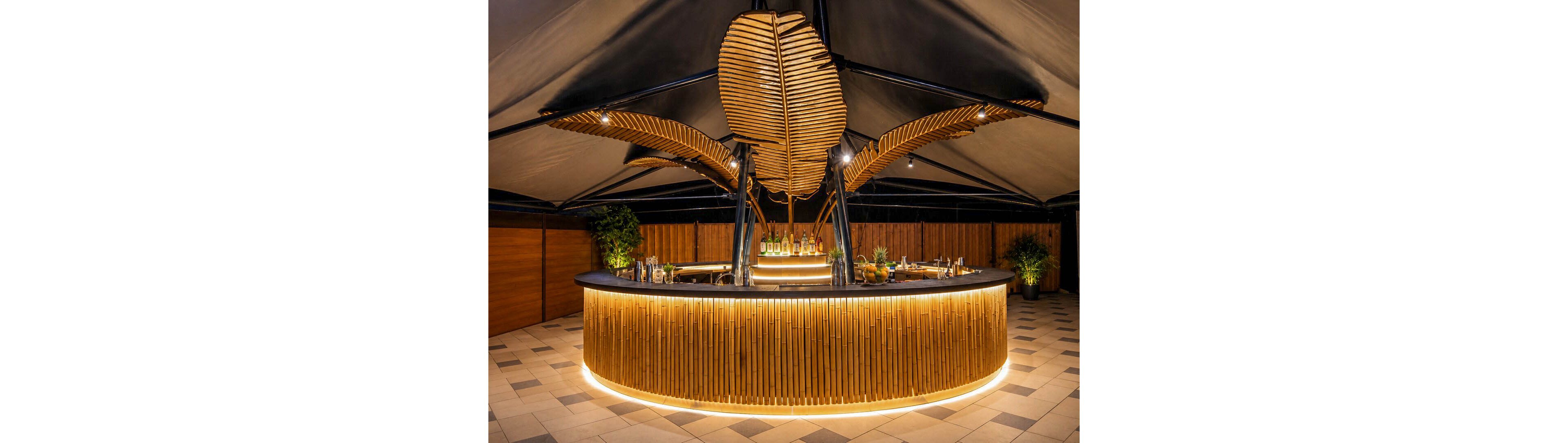 A stylish circular bar with warm wooden slats and integrated lighting stands in the center of a covered outdoor space. The bar counter displays various bottles, glassware, and cocktail-making tools. Large sculptural leaf-like structures rise above the bar, creating a tropical, contemporary focal point. Soft ambient lighting and potted plants enhance the inviting, upscale atmosphere.