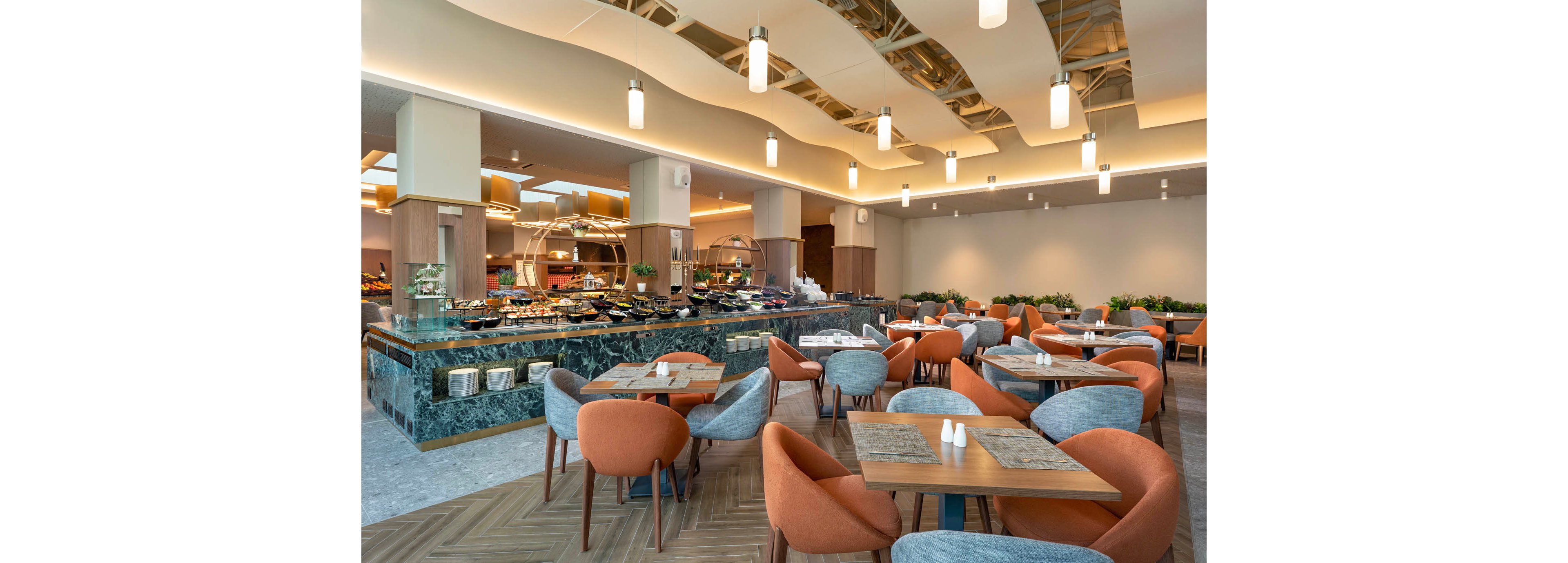 A contemporary restaurant interior features a spacious buffet area and neatly arranged dining tables. Orange and gray upholstered chairs surround wooden tables set with placemats and condiments. The buffet counter displays various dishes under warm lighting and decorative ceiling panels. Soft neutral tones, clean lines, and ample lighting create an inviting and modern dining atmosphere.