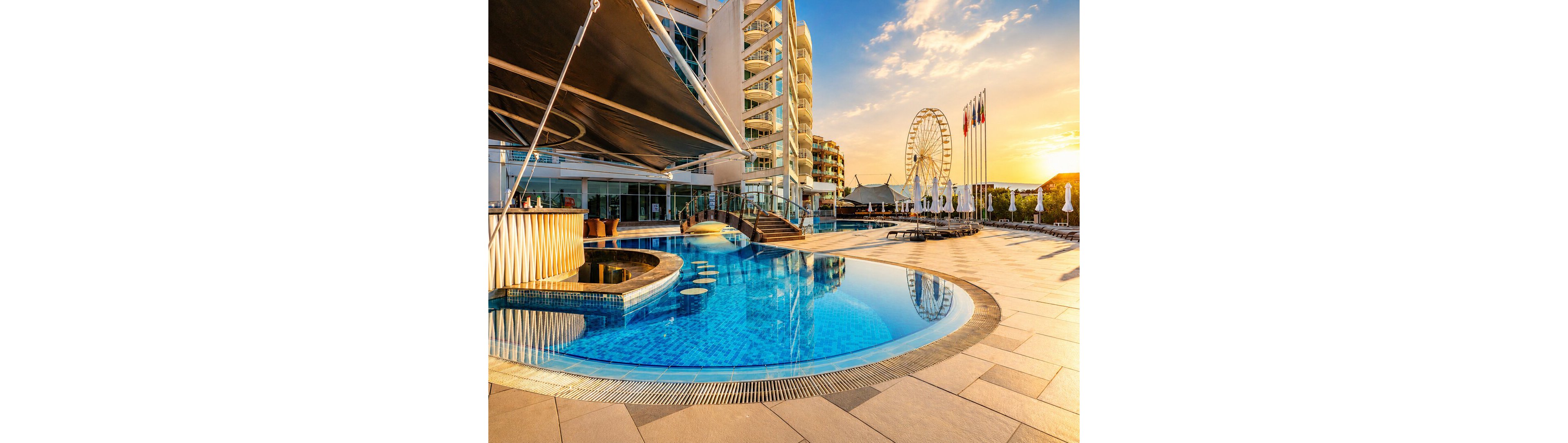 A luxurious resort swimming pool curves around a modern poolside bar at sunset. The scene features a contemporary hotel building with balconies overlooking the water. In the background, a large ferris wheel and multiple flagpoles add visual interest to the waterfront promenade. Warm golden light reflects off the calm blue pool, creating a relaxed vacation atmosphere.