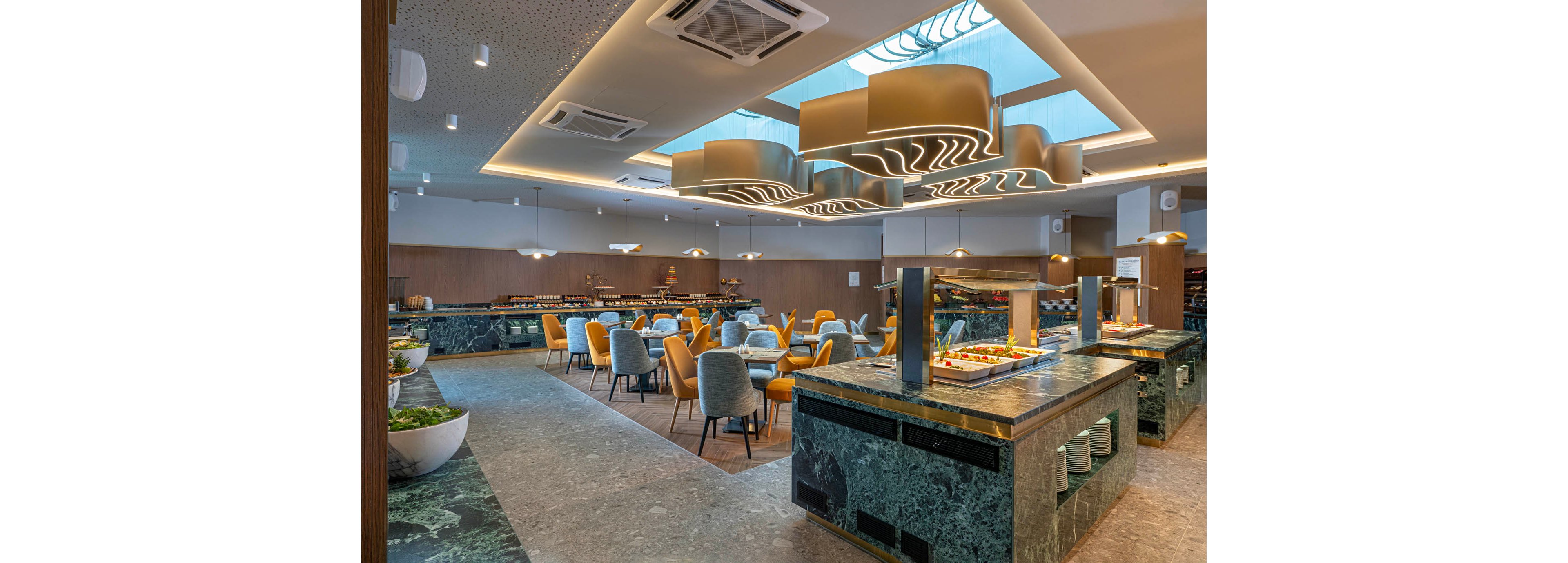 A contemporary buffet restaurant features marble counters displaying a variety of dishes. The space includes neatly arranged tables with upholstered chairs in yellow and gray tones. Sculptural ceiling lights and recessed lighting create a bright, inviting atmosphere. The overall design combines green stone surfaces, wood paneling, and a clean, modern aesthetic.