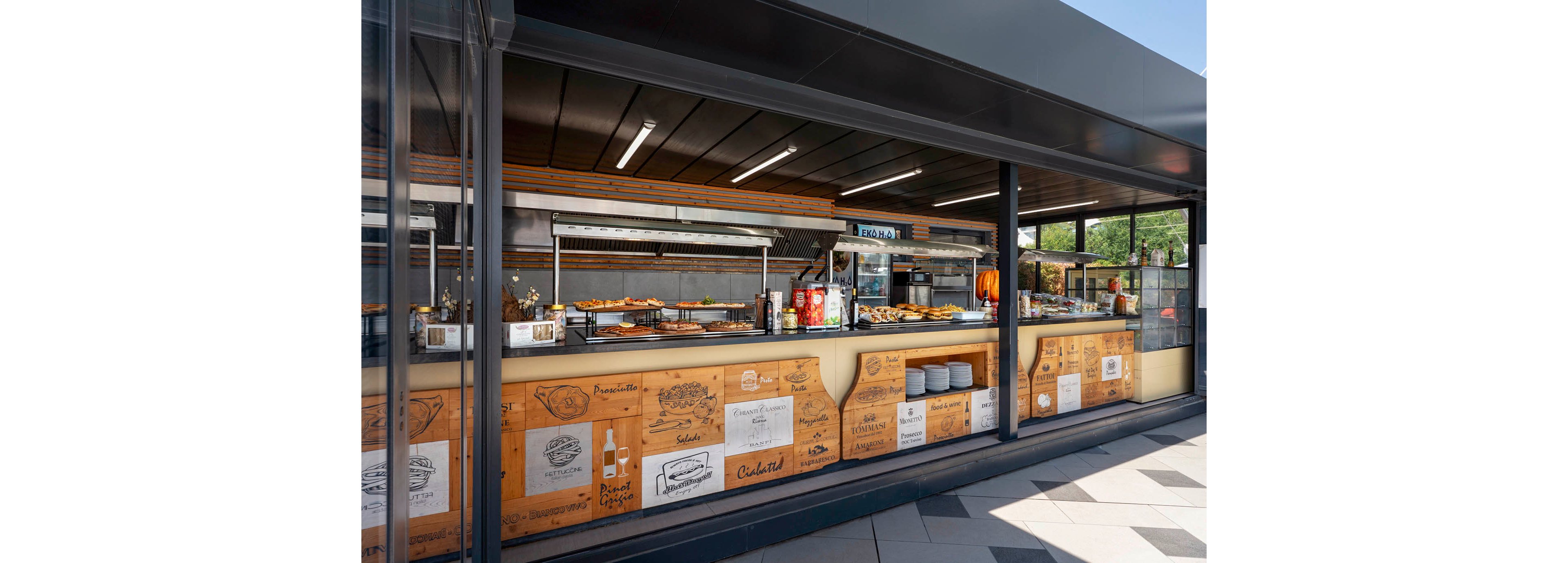 An open-air cafeteria counter showcases a variety of prepared foods behind glass. The setting appears to be a modern food court or casual dining area with a large serving line and stainless steel equipment. Illustrated panels with drawings of dishes and beverages decorate the wooden front of the counter. Bright daylight and clean geometric floor tiles contribute to a contemporary, inviting atmosphere.