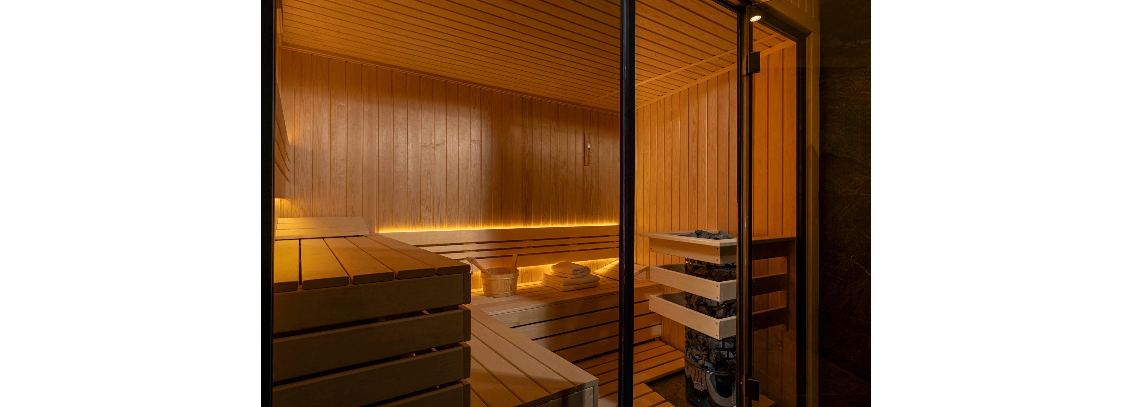 A contemporary wooden sauna interior is viewed through a glass door. Smooth horizontal wood paneling covers the walls, benches, and ceiling, creating a cohesive warm-toned space. Soft strip lighting runs along the back wall, highlighting neatly arranged towels and a wooden bucket. A heater filled with sauna stones stands in the corner, emphasizing the relaxing spa environment.
