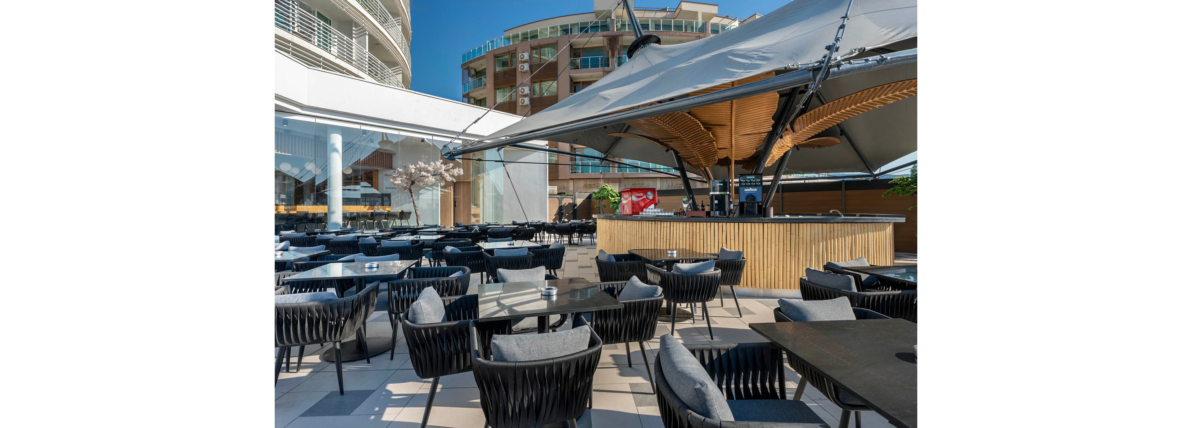 A contemporary open-air bar is set on a rooftop terrace with numerous black woven chairs and dark tables neatly arranged. The central bar structure features a wooden counter and a large tensile canopy providing shade. Surrounding buildings and blue sky indicate an urban setting with pleasant weather. The scene is empty of people, emphasizing the clean, modern design and relaxed atmosphere.