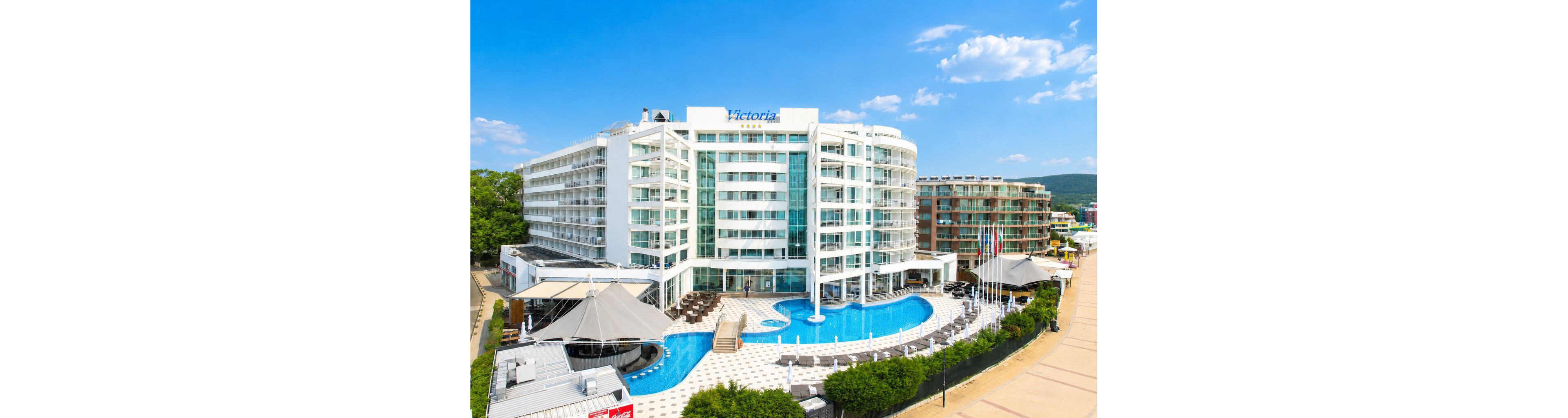 A large contemporary hotel with white facades and glass balconies stands prominently near a seaside promenade. The building features multiple floors arranged in a curved shape around a blue outdoor swimming pool. Sun loungers, umbrellas, and a patterned pool deck create a resort atmosphere. Nearby low-rise buildings and a clear blue sky suggest a sunny coastal vacation destination.