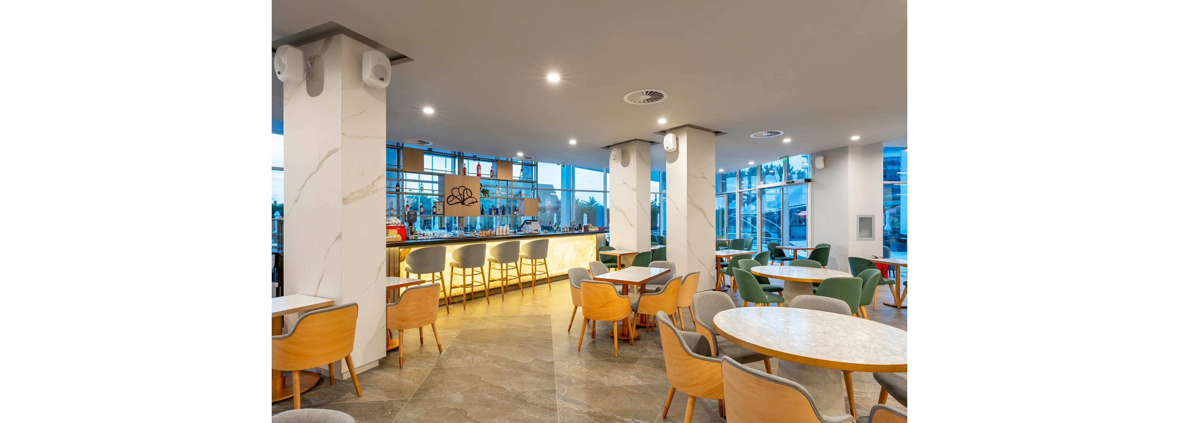 A modern hotel bar interior features a long illuminated counter with high stools and a variety of colorful bottles displayed on glass shelves. In the foreground, round tables with mixed wooden and upholstered chairs create a casual seating area. Large windows allow natural light to fill the space and offer views of the outdoors. Neutral tones, clean lines, and subtle lighting contribute to a calm, contemporary atmosphere.