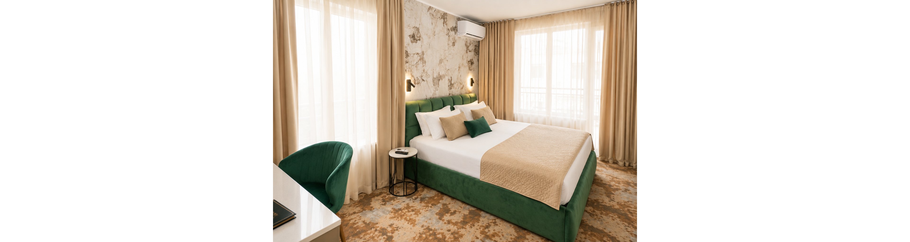 A contemporary hotel bedroom features a large green upholstered bed neatly made with beige bedding and accent pillows. The room is bright, with natural light streaming through tall windows covered by sheer and beige curtains. A patterned carpet, marble-effect accent wall, and minimalist wall sconces add a stylish touch. A small round side table, desk, and green chair complete the cozy, inviting space.
