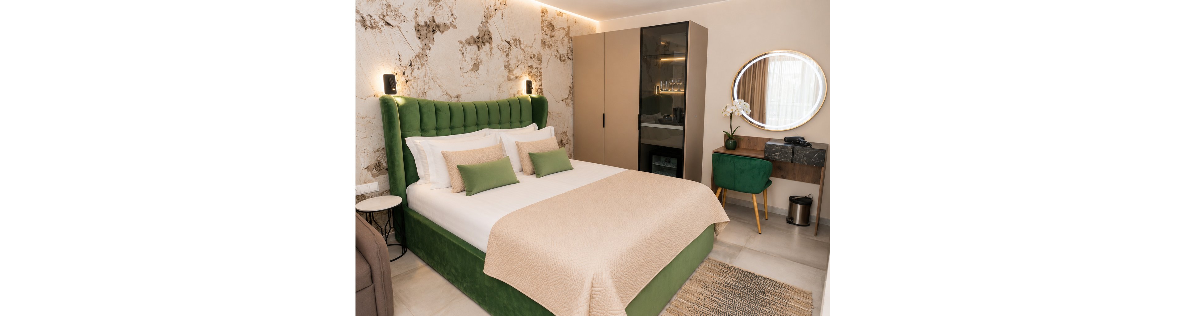 A contemporary bedroom features a large bed with a green upholstered frame and neutral bedding. The room includes a sleek wardrobe with reflective doors and a small vanity area with a round mirror. A green chair and potted orchid add color and elegance to the space. Soft wall lighting and a marble-patterned accent wall create a warm, inviting atmosphere.