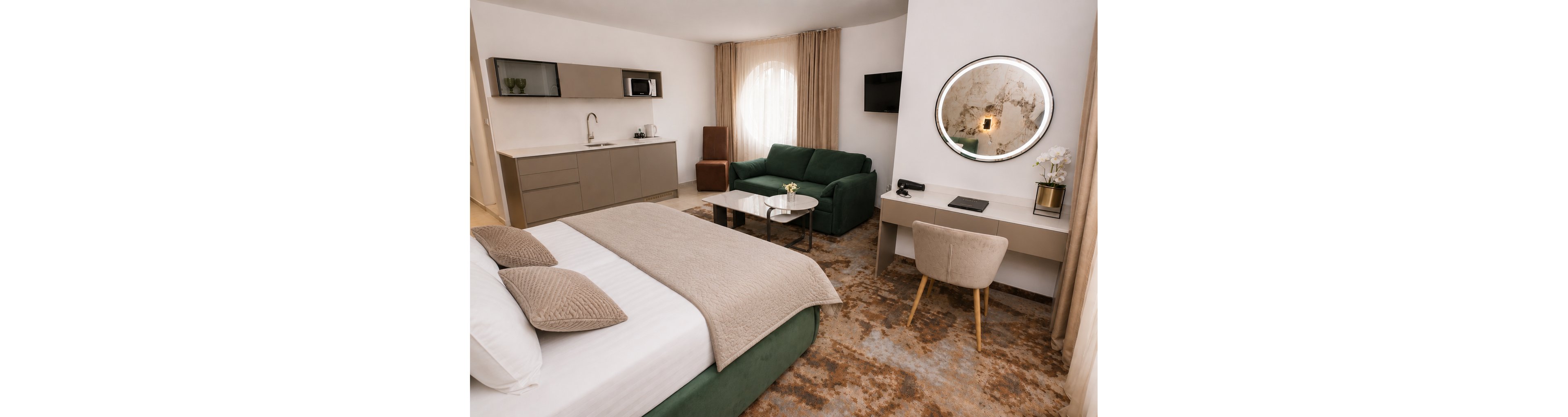A contemporary hotel suite features a neatly made double bed with neutral bedding in the foreground. In the background, a green sofa and coffee tables sit near a large round window with sheer curtains. A compact kitchenette with cabinets and a sink lines one wall, while a desk with a chair, mirror, and small television creates a functional workspace. Warm beige tones, soft lighting, and a patterned carpet contribute to a cozy, modern atmosphere.