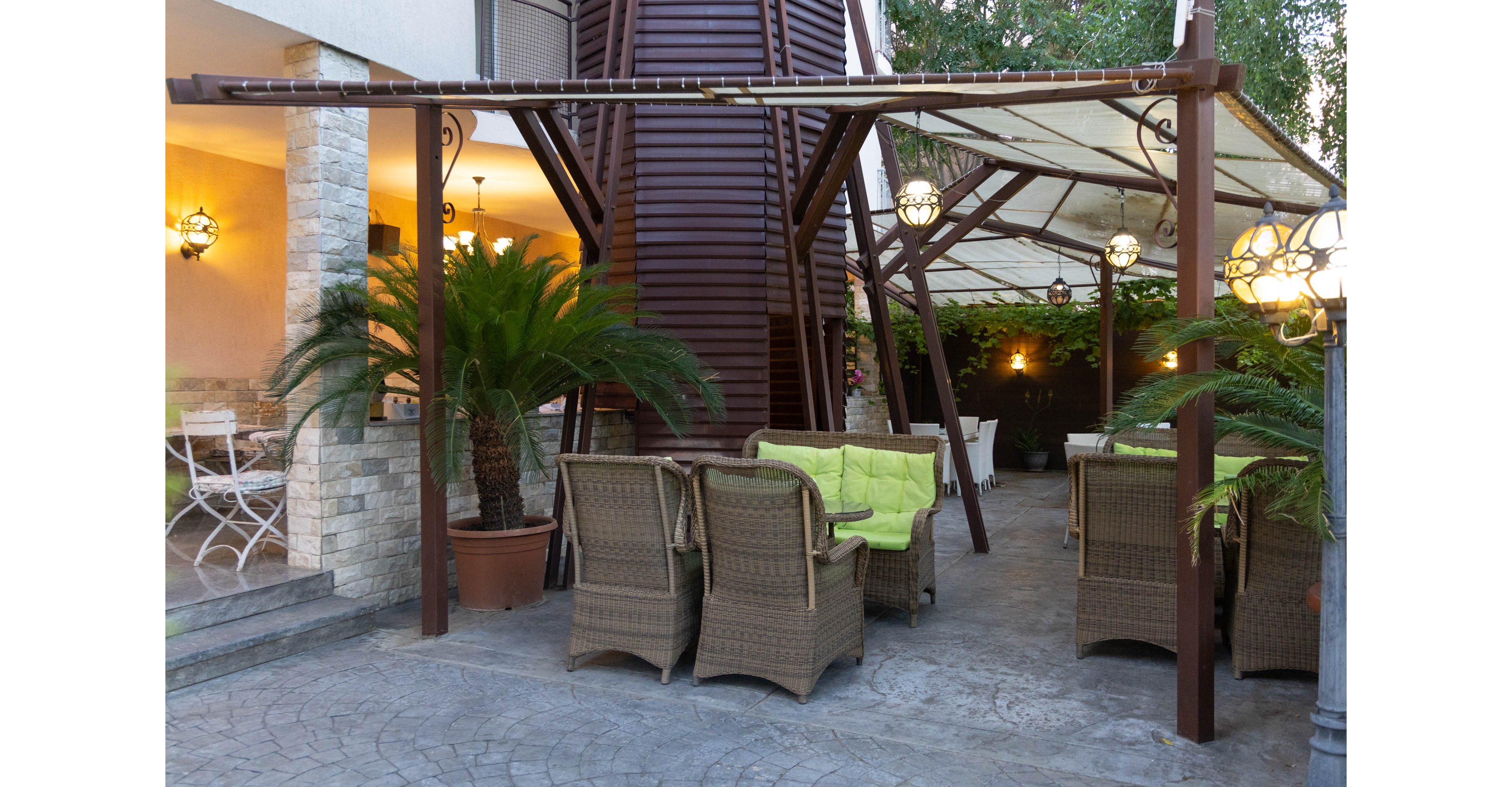 An inviting outdoor patio features wicker armchairs and sofas arranged around a small table. The seating area is covered by a pergola-style canopy with warm hanging lanterns providing ambient light. Potted palm plants and lush greenery line the space, adding a tropical touch to the stone flooring and walls. The overall setting suggests a comfortable place for relaxation or social gatherings in the evening.