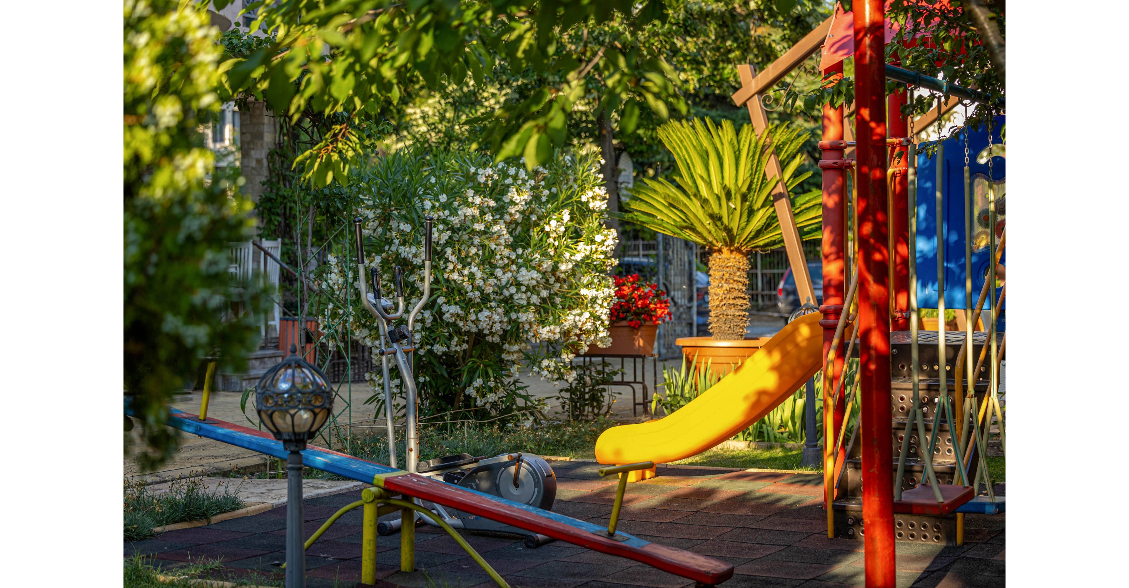A small playground with bright equipment sits in a sunlit garden. A yellow slide and seesaw stand on rubber flooring surrounded by trees and flowering bushes. Warm evening light highlights red and blue metal structures and a potted palm. The scene feels quiet and inviting, suggesting a peaceful neighborhood play area.