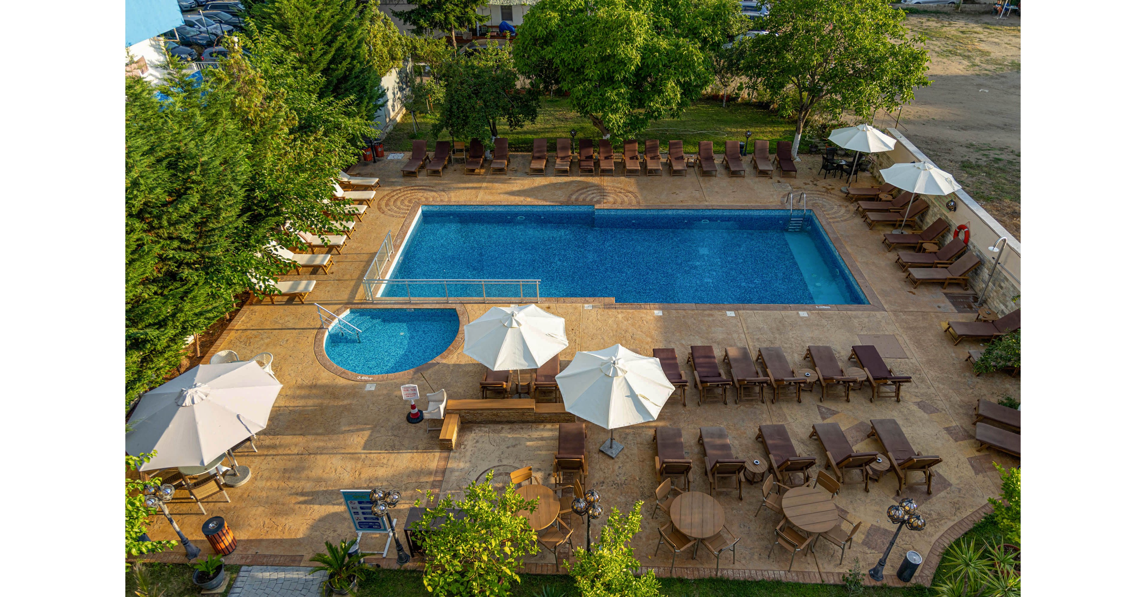 A rectangular outdoor swimming pool is surrounded by numerous sun loungers and parasols on a tiled terrace. A smaller circular pool sits adjacent to the main pool, separated by a low barrier. Trees and greenery frame the pool area, with parked cars and open ground visible beyond the fence. The scene is brightly lit, suggesting a warm, sunny day at a resort or hotel.