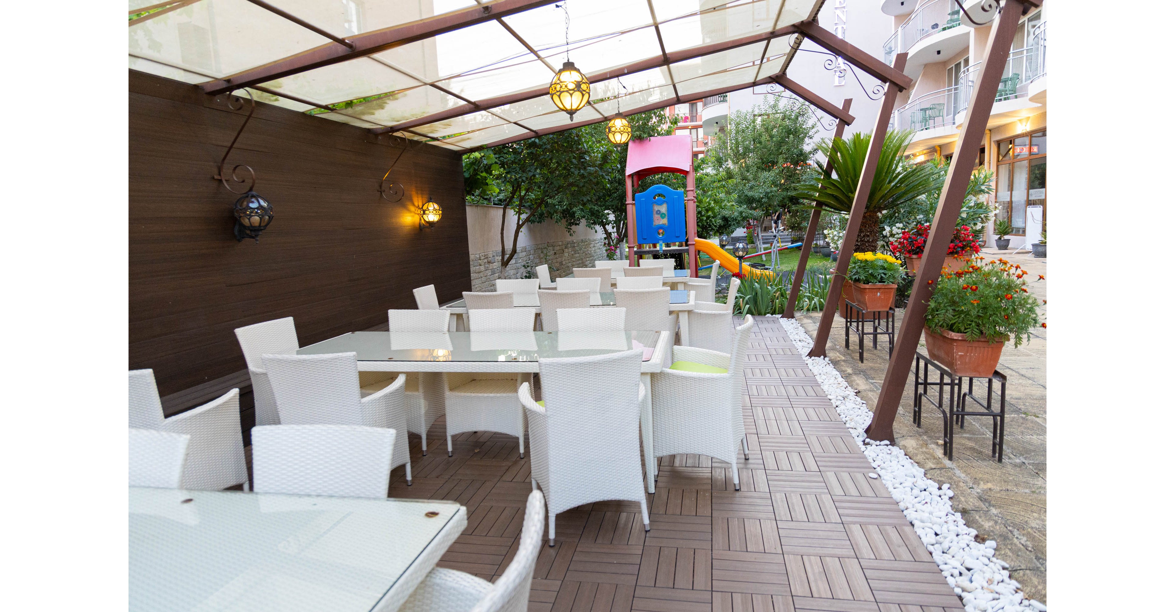 An outdoor cafe terrace features white wicker chairs and glass-top tables arranged on a patterned wooden deck. Decorative hanging lights and a translucent canopy cover the seating area, creating a cozy atmosphere. To the side, potted red flowers and lush greenery add color and texture. In the background, a bright blue and orange playground slide stands near residential buildings, suggesting a family-friendly setting.