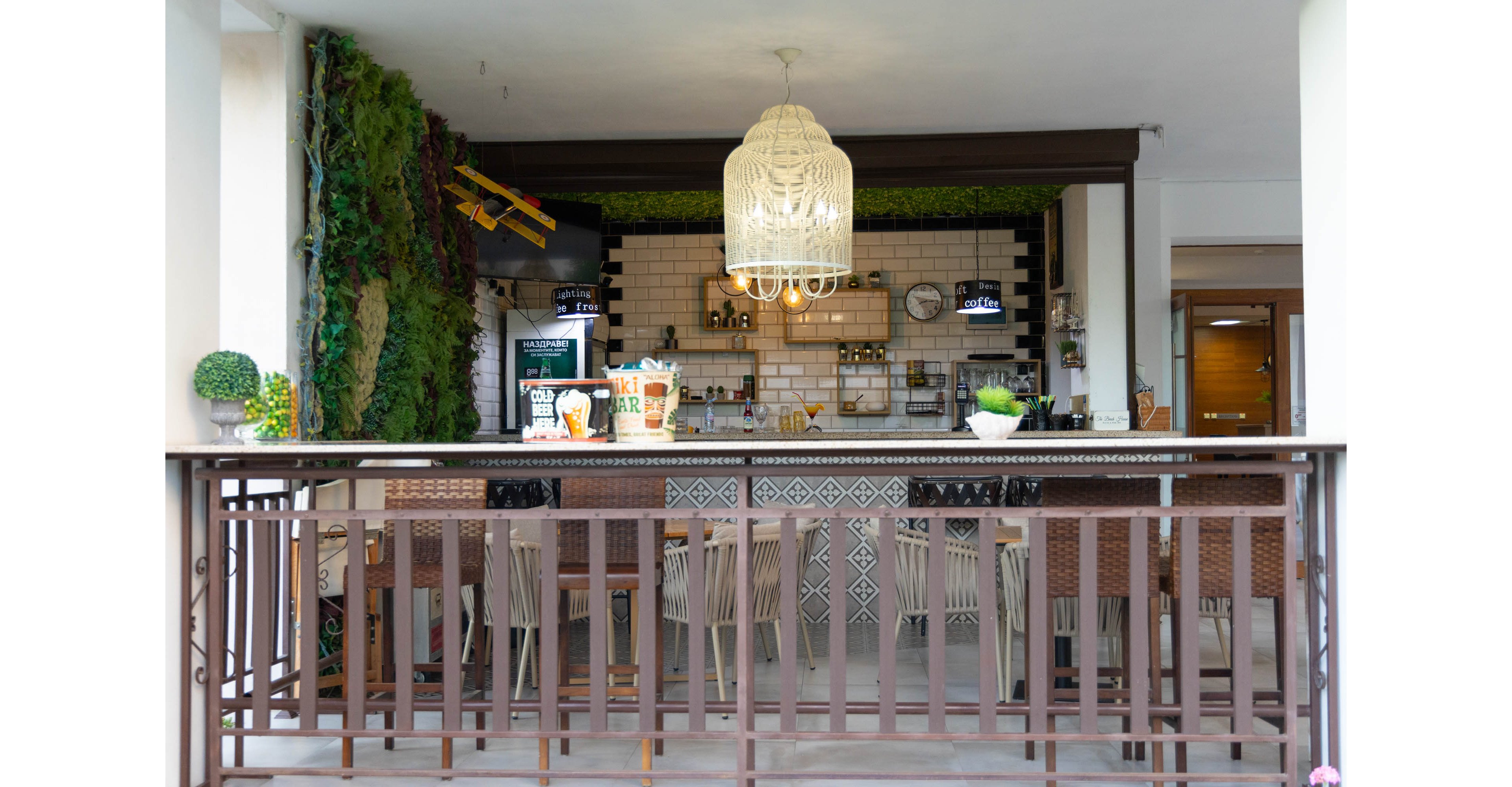 An inviting bar counter is set in an open-air space with a decorative railing in the foreground. The back wall features white subway tiles, shelves with bottles and glassware, and warm pendant lights. A large white woven chandelier hangs above the bar, while greenery and a small model airplane add visual interest on the side wall. Wooden stools line the counter, creating a relaxed, casual atmosphere suitable for social gatherings.