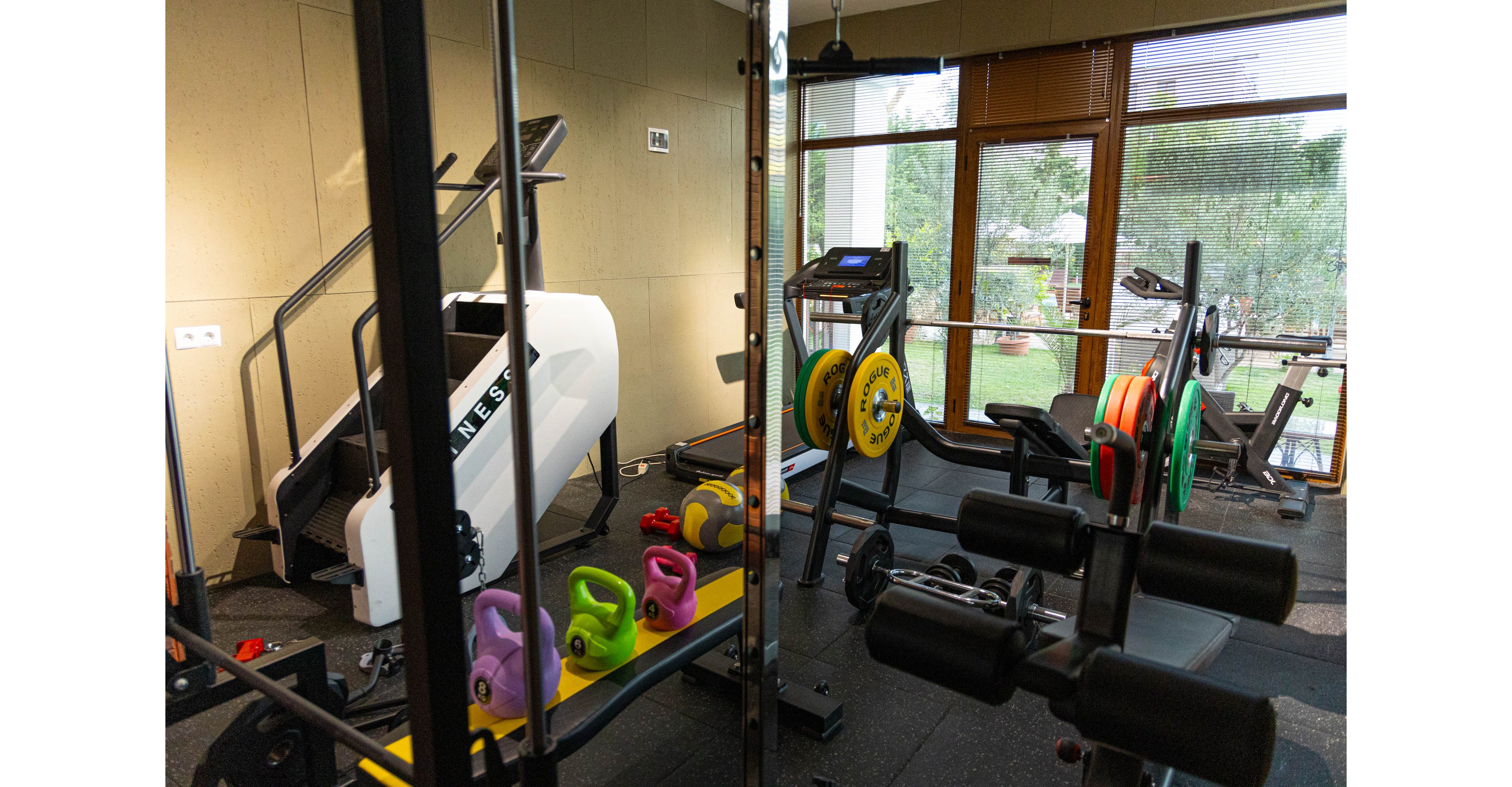A well-equipped home gym features various strength training machines and free weights. Colorful kettlebells sit on a bench alongside barbells loaded with weight plates. Large windows reveal a green outdoor garden, allowing natural light to fill the workout space. The room has a clean, organized appearance with rubber flooring and contemporary fitness equipment.