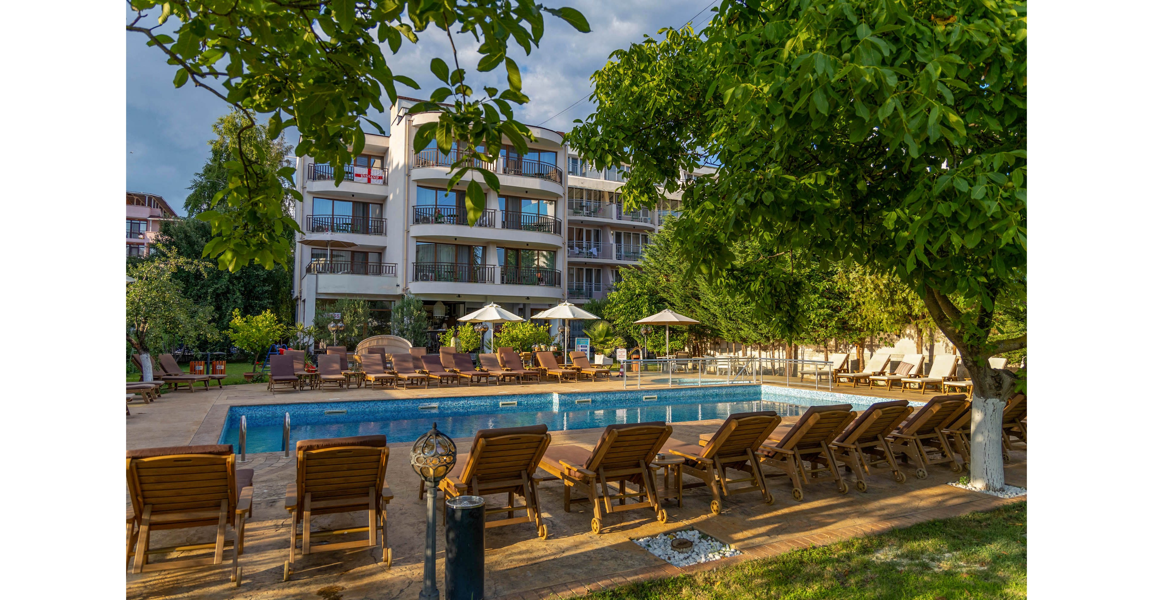 An outdoor swimming pool is surrounded by wooden lounge chairs in front of a modern multi-story hotel building. White umbrellas and seating areas line the poolside, creating a relaxed vacation atmosphere. Lush green trees and landscaped gardens frame the scene, adding natural shade and color. Warm sunlight enhances the inviting resort setting and highlights the architectural details of the building.