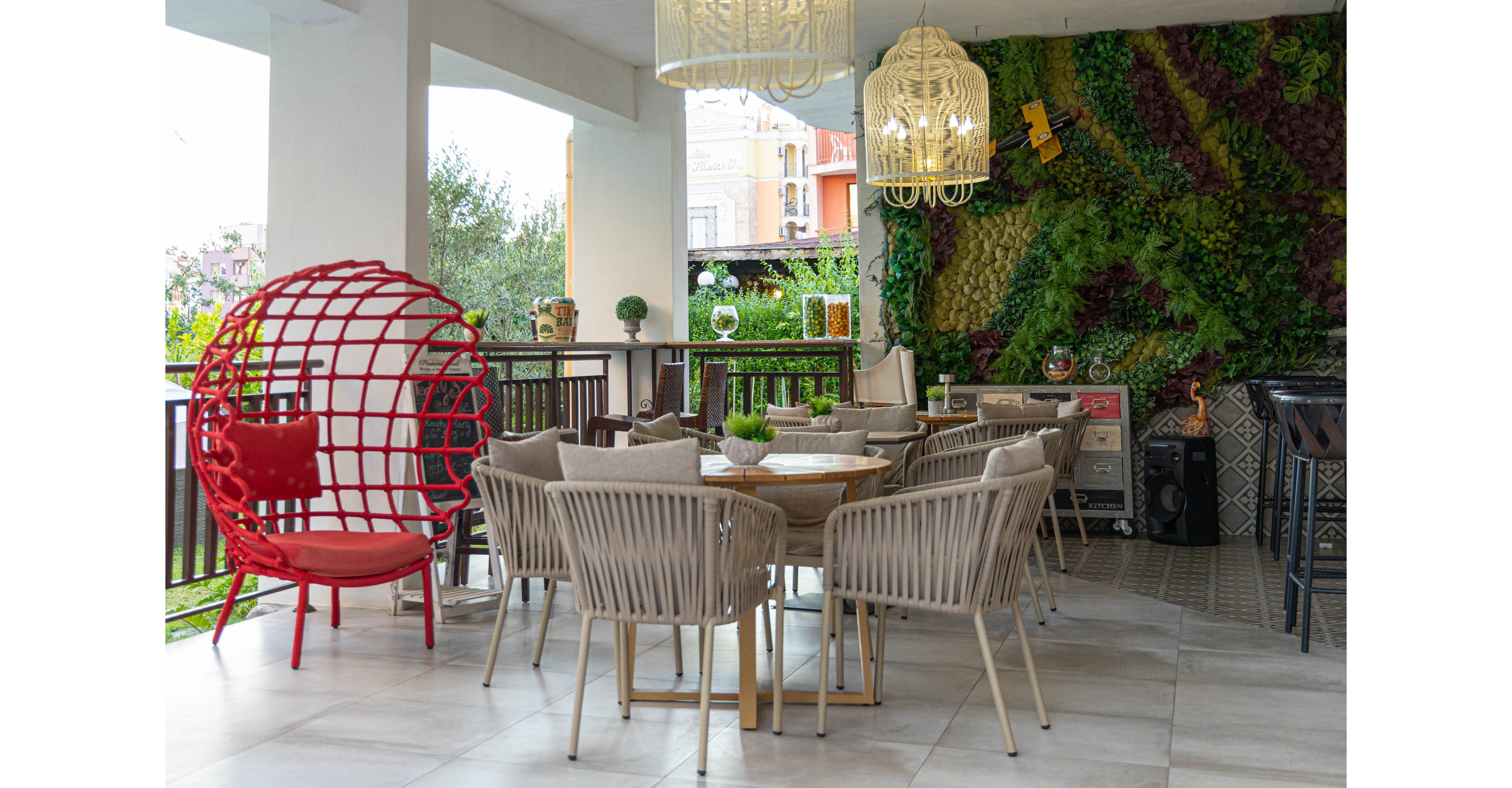 Contemporary outdoor seating area features a round wooden table surrounded by beige woven chairs. A striking red cage-style chair stands to the side, adding a bold accent to the neutral decor. The space is framed by a lush vertical garden wall and decorative hanging light fixtures. Natural light and garden views create a relaxed, inviting atmosphere suitable for dining or social gatherings.