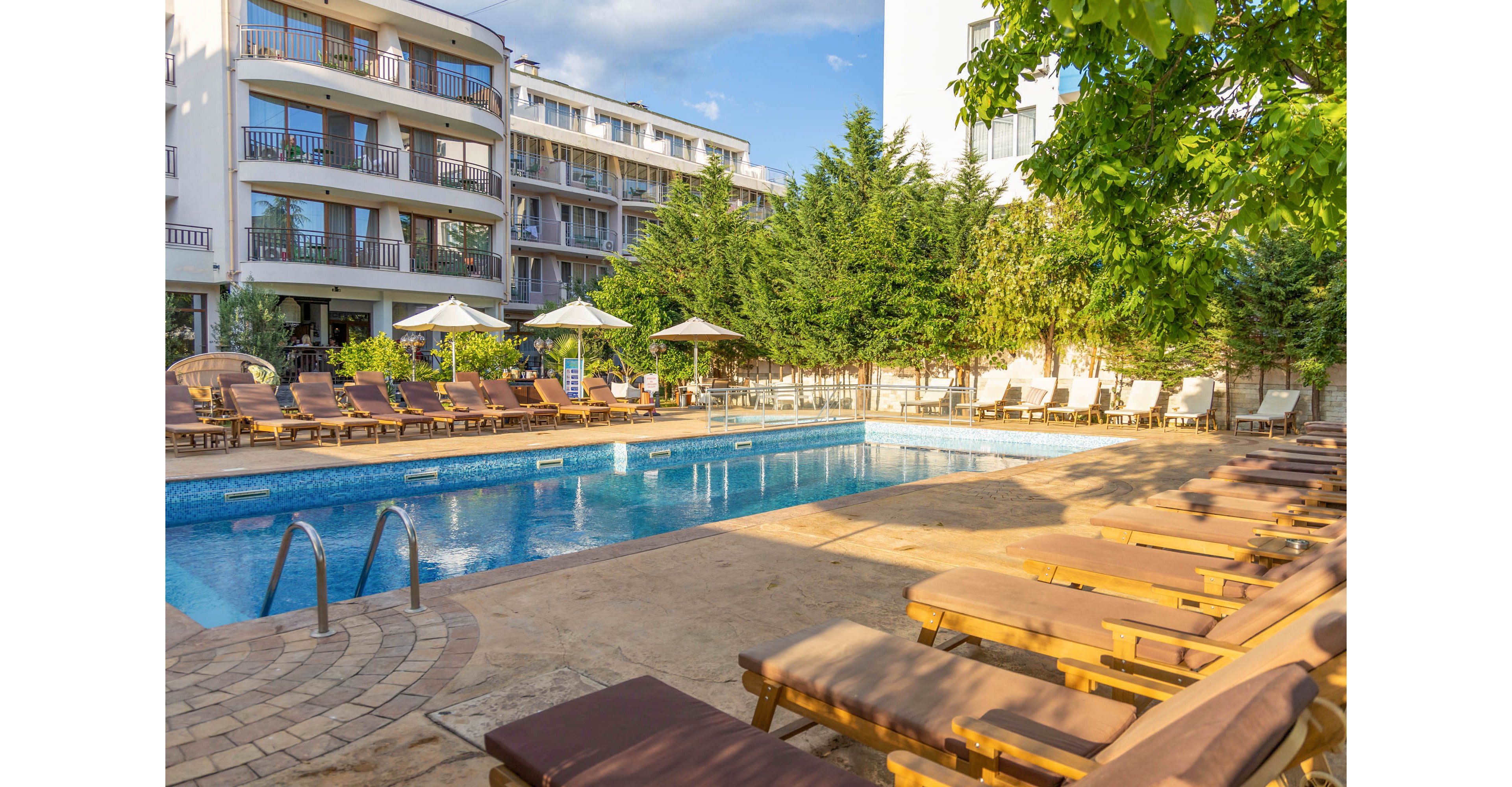 An outdoor swimming pool is surrounded by wooden lounge chairs and umbrellas in a hotel courtyard. Multi-story apartment-style buildings frame the pool area, with balconies overlooking the scene. Lush green trees and shrubs add a natural, relaxing atmosphere. The warm sunlight and clear sky suggest a pleasant summer day at a resort.