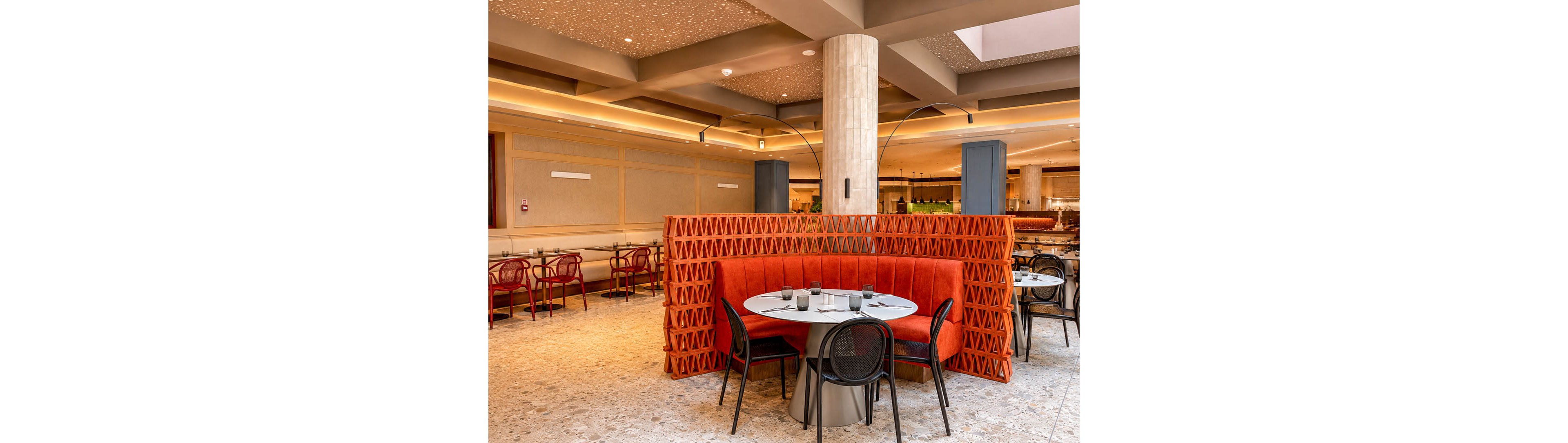 A contemporary restaurant interior features a round table set within a curved orange booth. The seating area is framed by a decorative geometric partition and surrounded by additional black chairs and tables. Warm ceiling lights, terrazzo flooring, and neutral wall panels create a cozy yet upscale atmosphere. The space appears ready for guests, with glassware neatly arranged on the tables.