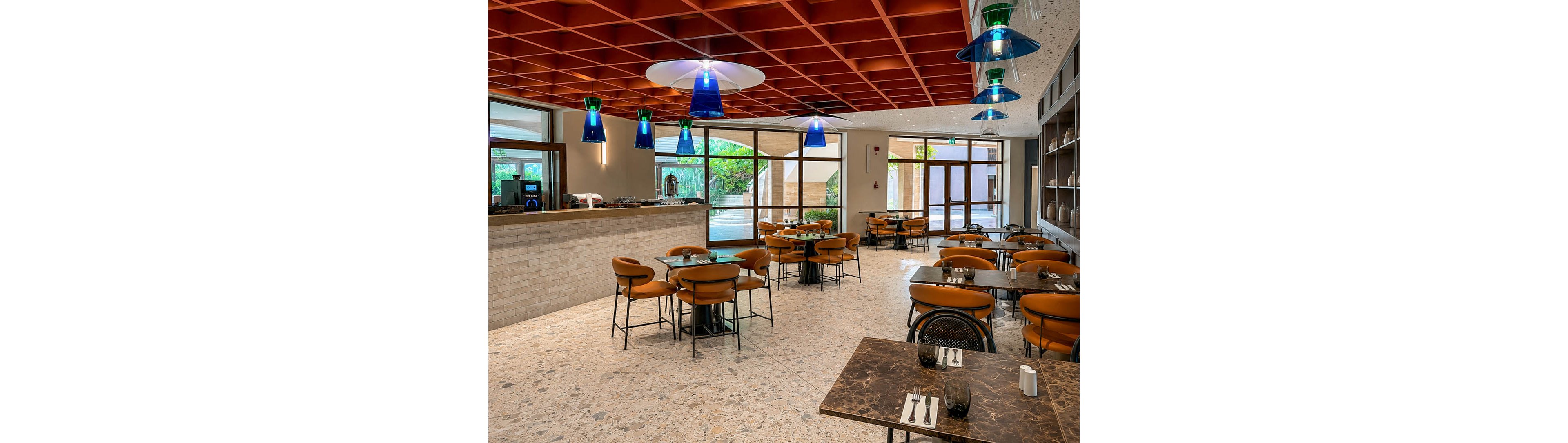 A contemporary cafe interior features neatly arranged tables and leather chairs around a stone-topped counter. Large windows and glass doors let in natural light and reveal greenery outside. Blue and green pendant lights hang from a geometric wooden ceiling, casting a warm glow over the terrazzo floor. The space is clean, organized, and ready to welcome guests, with cutlery and small decor items set on the tables.