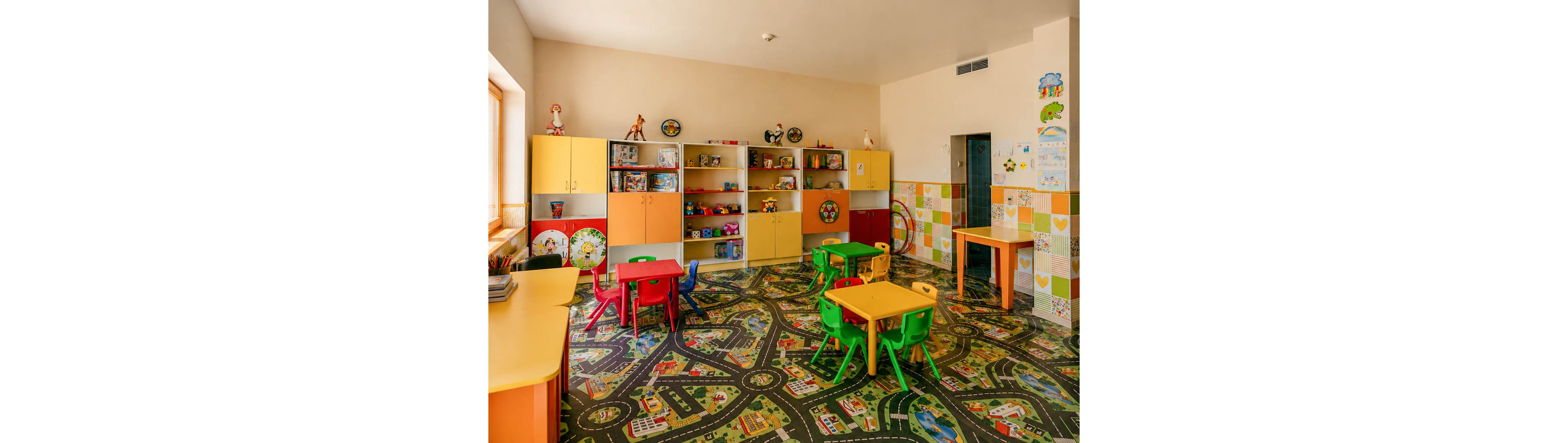 A bright kindergarten classroom features small colorful tables and chairs arranged on a patterned play carpet. The floor design shows roads, buildings, and greenery, creating a playful city map for children. Along the back wall, shelves and cabinets hold toys, books, and decorative items in various shapes and colors. Sunlight enters through a large window, enhancing the cheerful and inviting atmosphere of the room.