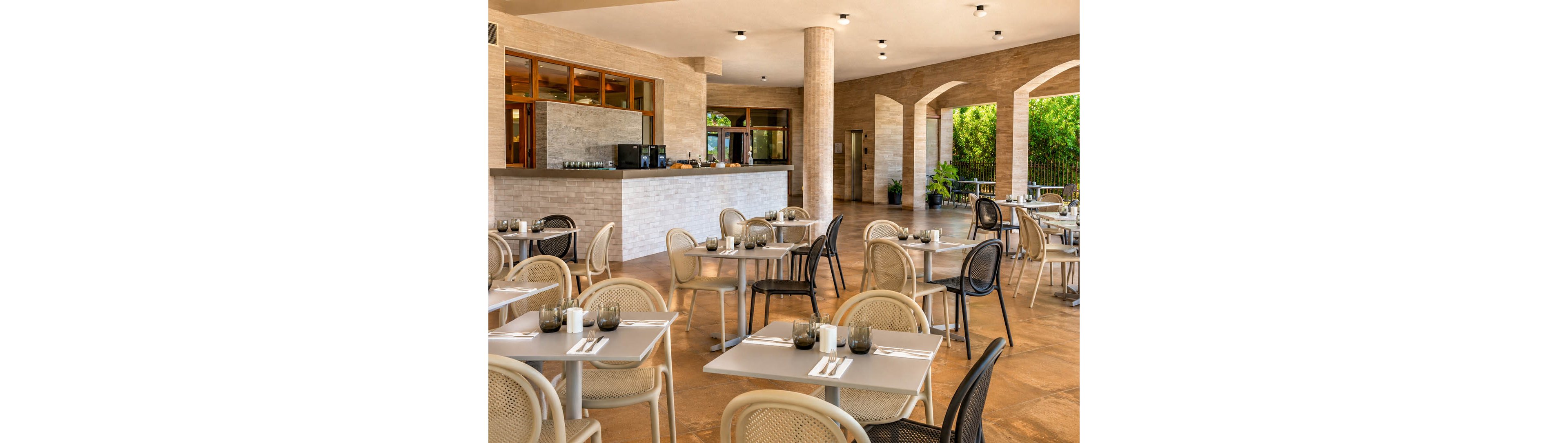 A contemporary open air restaurant features neatly arranged tables and chairs on a warm-toned floor. Each table is set with cutlery, napkins, and glasses, ready for guests. A minimalist bar counter with coffee equipment stands in the background beneath a covered terrace. Large arches open to a green outdoor area, creating a bright and inviting dining atmosphere.