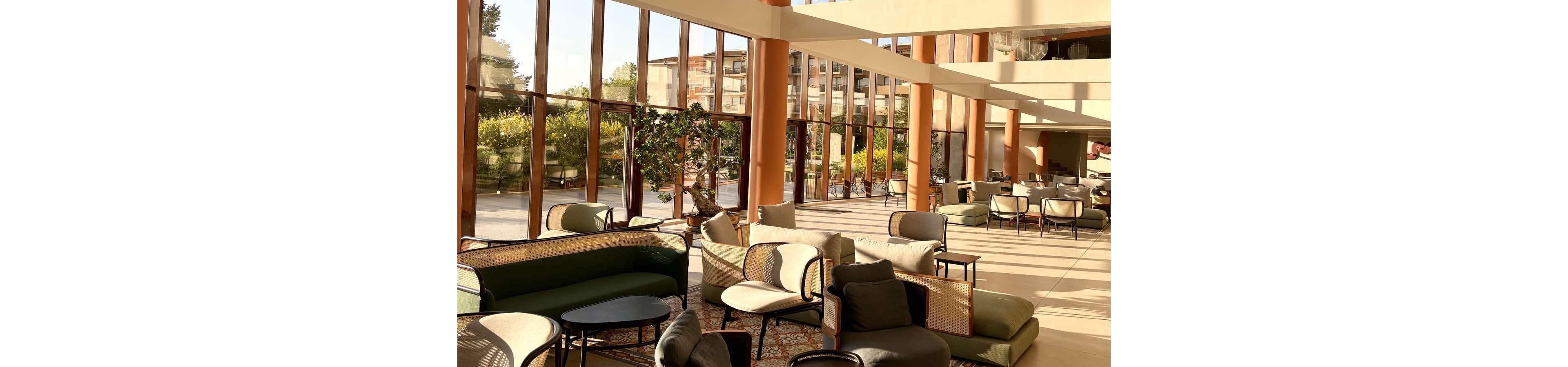 A spacious hotel lobby lounge is filled with contemporary sofas and armchairs arranged in conversational seating areas. Floor-to-ceiling windows let in abundant natural light and reveal greenery and buildings outside. Warm sunlight casts long shadows across the polished floor and soft neutral-toned furnishings. The overall atmosphere feels relaxed, inviting, and ideal for socializing or informal meetings.