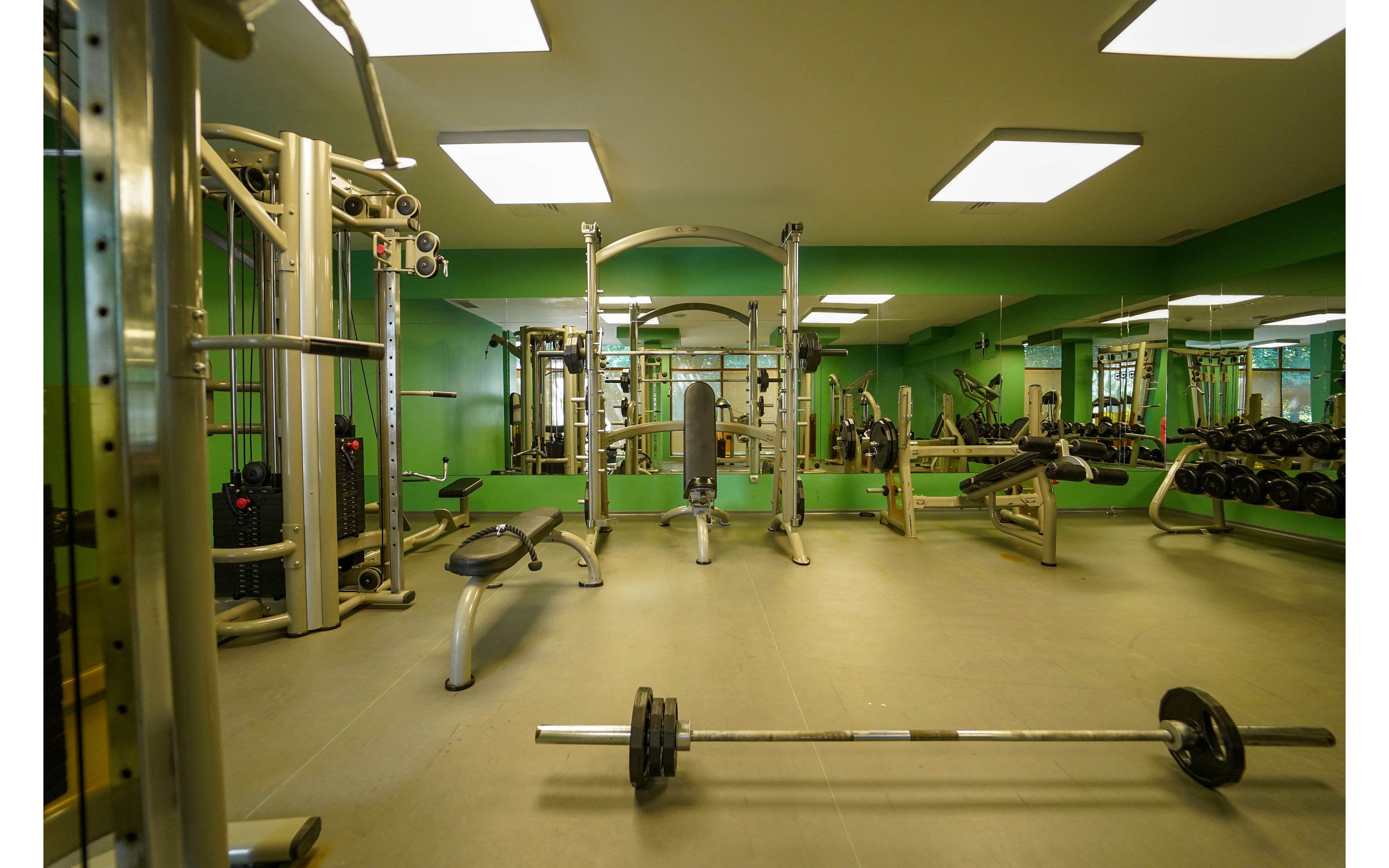 A clean, modern gym interior features various strength training machines and free weights. A barbell with weight plates rests on the floor in the foreground, while benches and cable machines line the room. Large mirrors on the walls reflect the equipment and create a sense of spaciousness. Bright ceiling lights and green wall accents give the fitness space an energetic, contemporary feel.