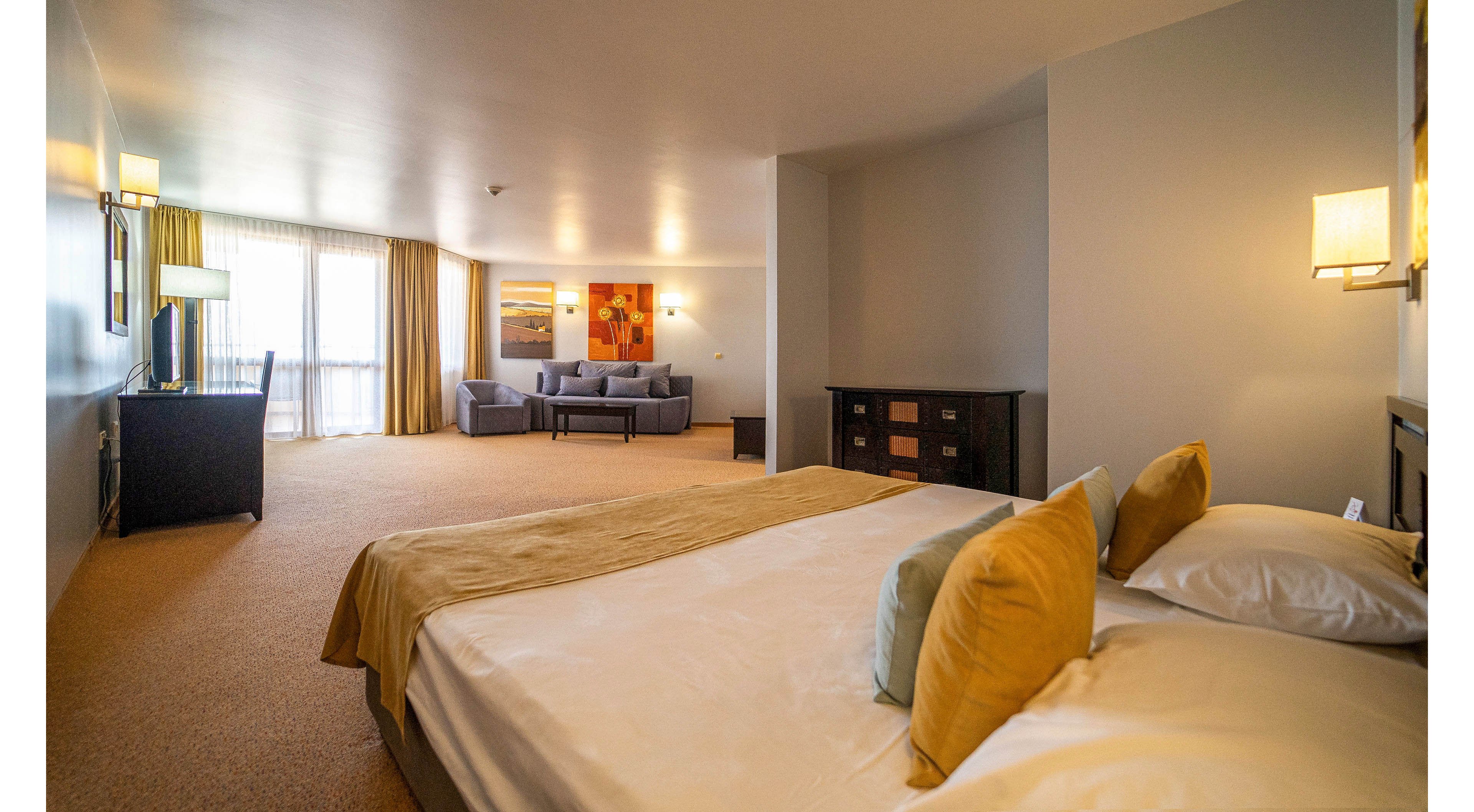 A large, modern hotel room features a neatly made double bed with neutral bedding and accent pillows in warm tones. In the background, a cozy seating area with a sofa and armchairs sits near wide windows covered by light curtains, allowing natural light to fill the space. Soft wall lighting and contemporary artwork add a welcoming, upscale atmosphere. The overall palette is warm and muted, emphasizing comfort and relaxation.