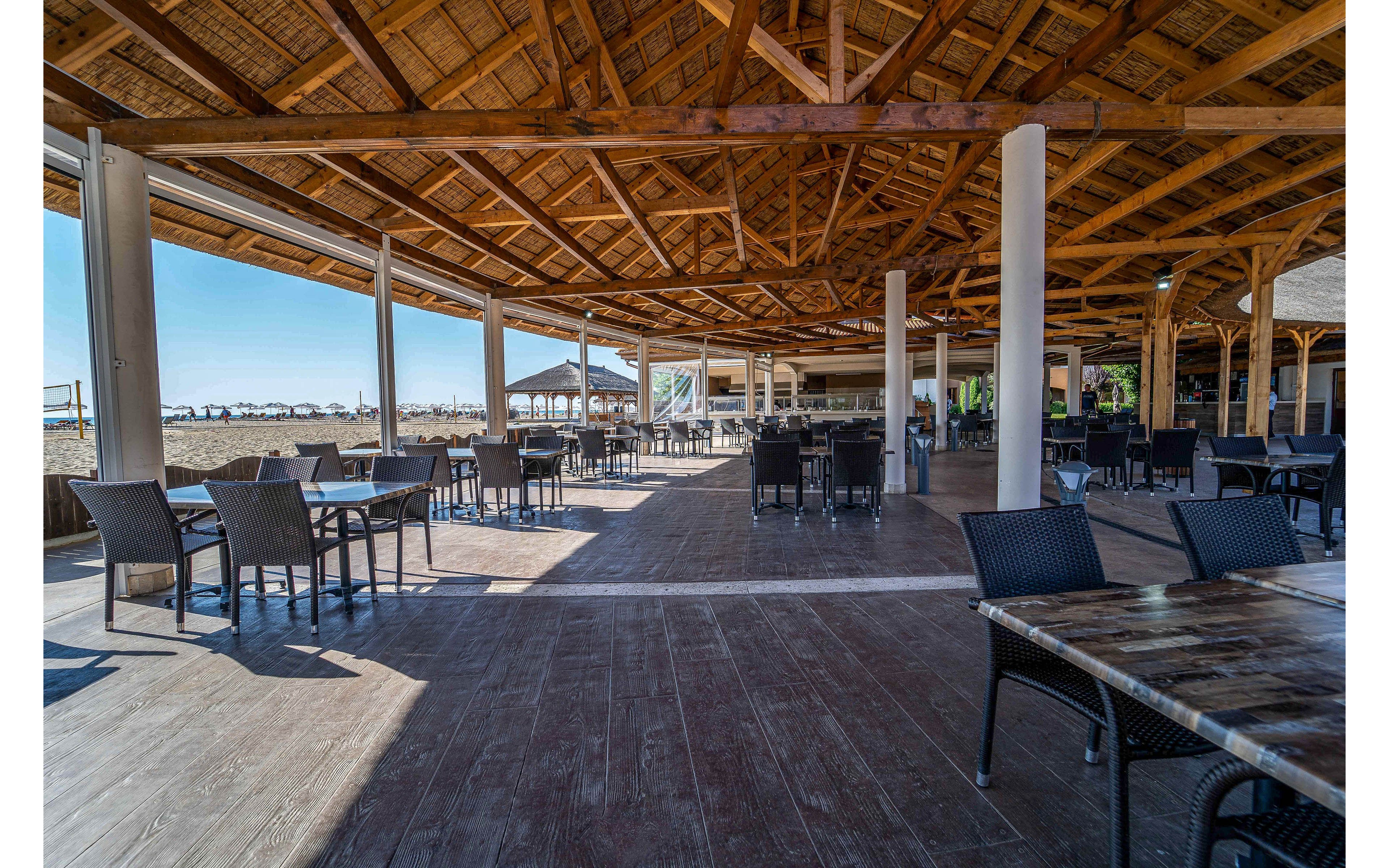 Spacious open-air restaurant with wicker chairs and tables sits under a large wooden thatched roof. The setting overlooks a sunny beachfront with umbrellas and lounge chairs visible in the distance. Strong daylight casts long shadows across the wooden floor, emphasizing the airy, coastal atmosphere. The scene appears calm and uncrowded, suggesting a relaxed seaside dining experience.