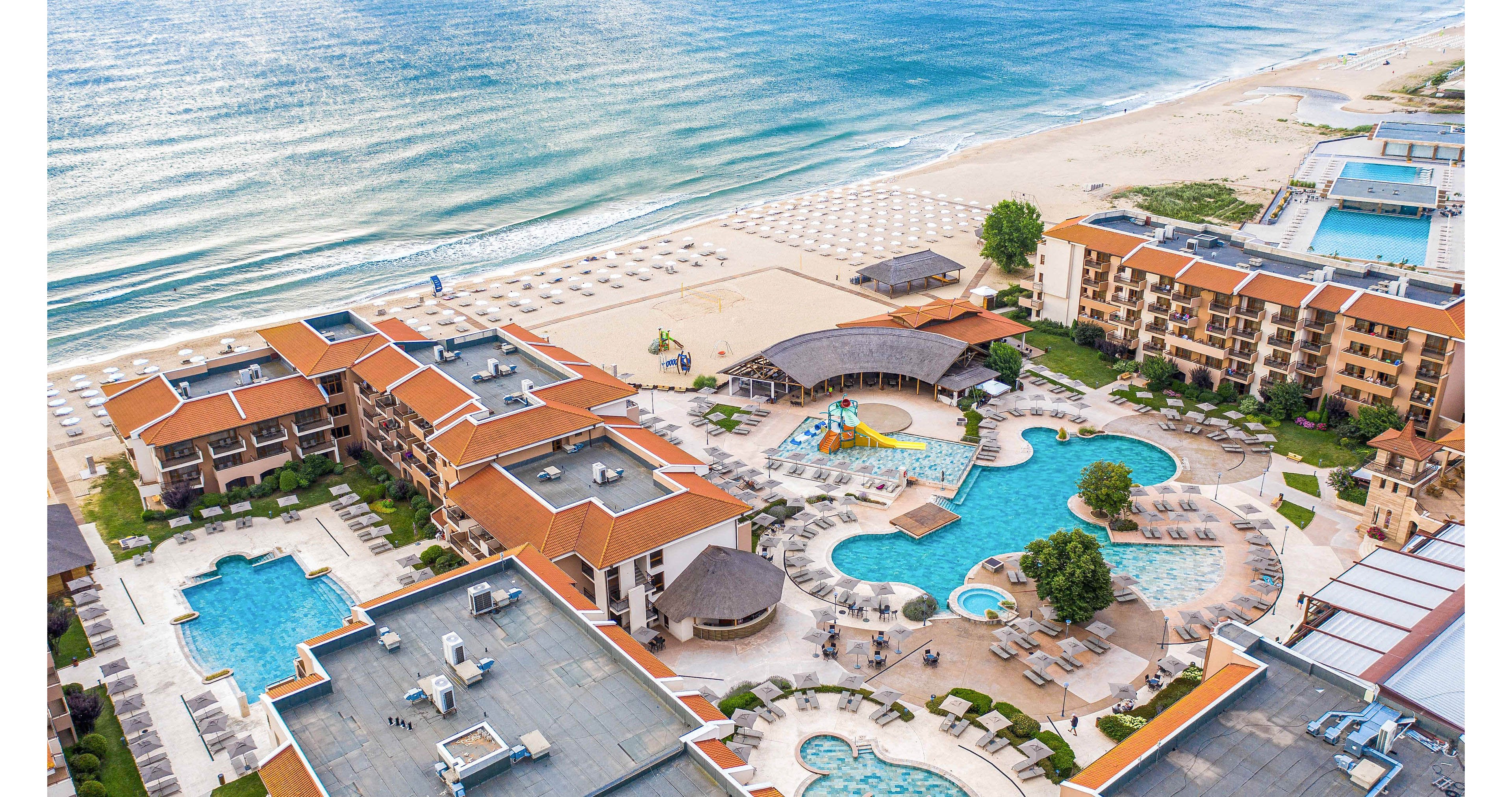 A large seaside resort complex is captured from above, showing multiple buildings with red roofs surrounding several outdoor swimming pools. The property sits directly on a sandy beach that meets calm ocean waves. Sun loungers, umbrellas, and landscaped pathways create a relaxed vacation atmosphere around the pools. The overall scene suggests a warm coastal destination ideal for holidays and leisure.