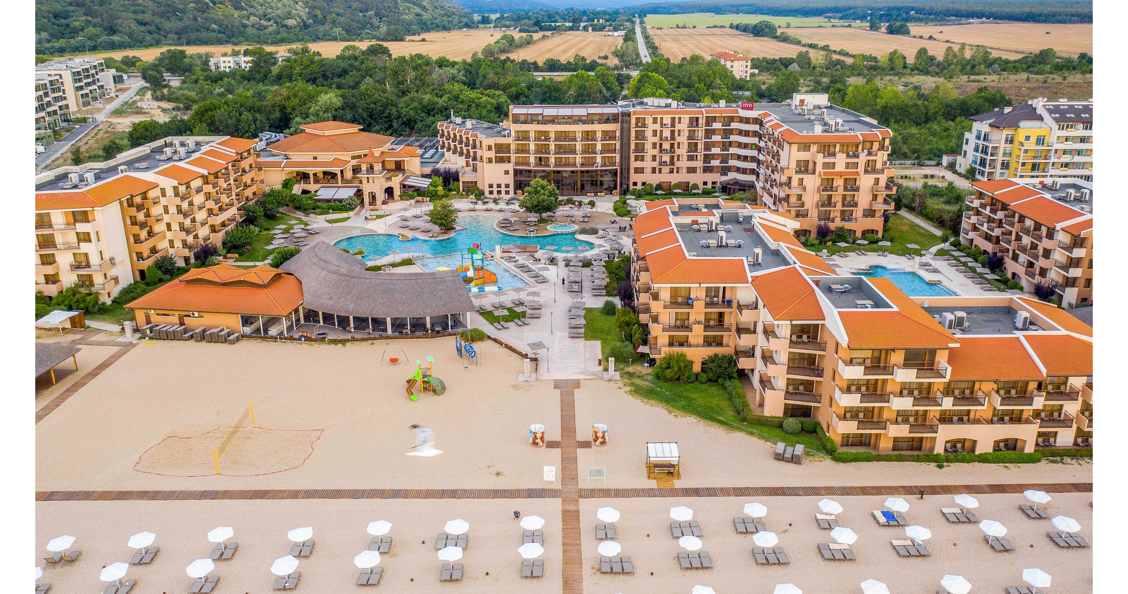 A large seaside resort complex with multiple tan and orange-roofed buildings is captured from an aerial perspective. The property features several outdoor swimming pools, a central courtyard, and a landscaped area surrounded by trees and fields in the distance. In the foreground, a wide sandy beach with rows of white umbrellas and sun loungers stretches toward the water. A volleyball court and small playground structures are visible on the sand, emphasizing leisure and family activities.