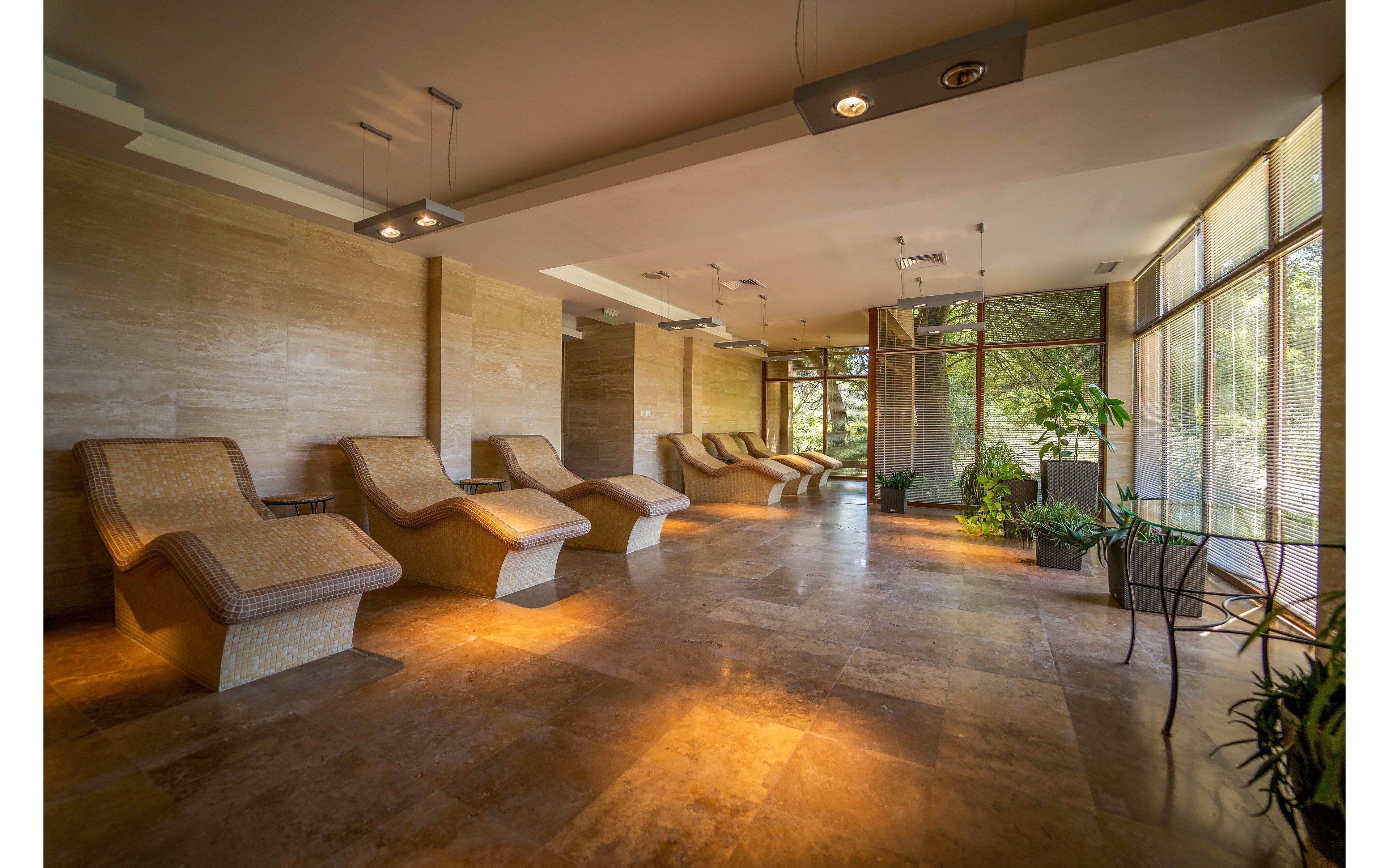 A spacious spa relaxation lounge features several curved reclining chairs arranged along a warm-toned stone wall. Large floor-to-ceiling windows on one side let in natural light and offer views of greenery outside. Potted plants are placed throughout the room, softening the contemporary interior design. Recessed and pendant lighting create a calm, inviting atmosphere suitable for rest and wellness.