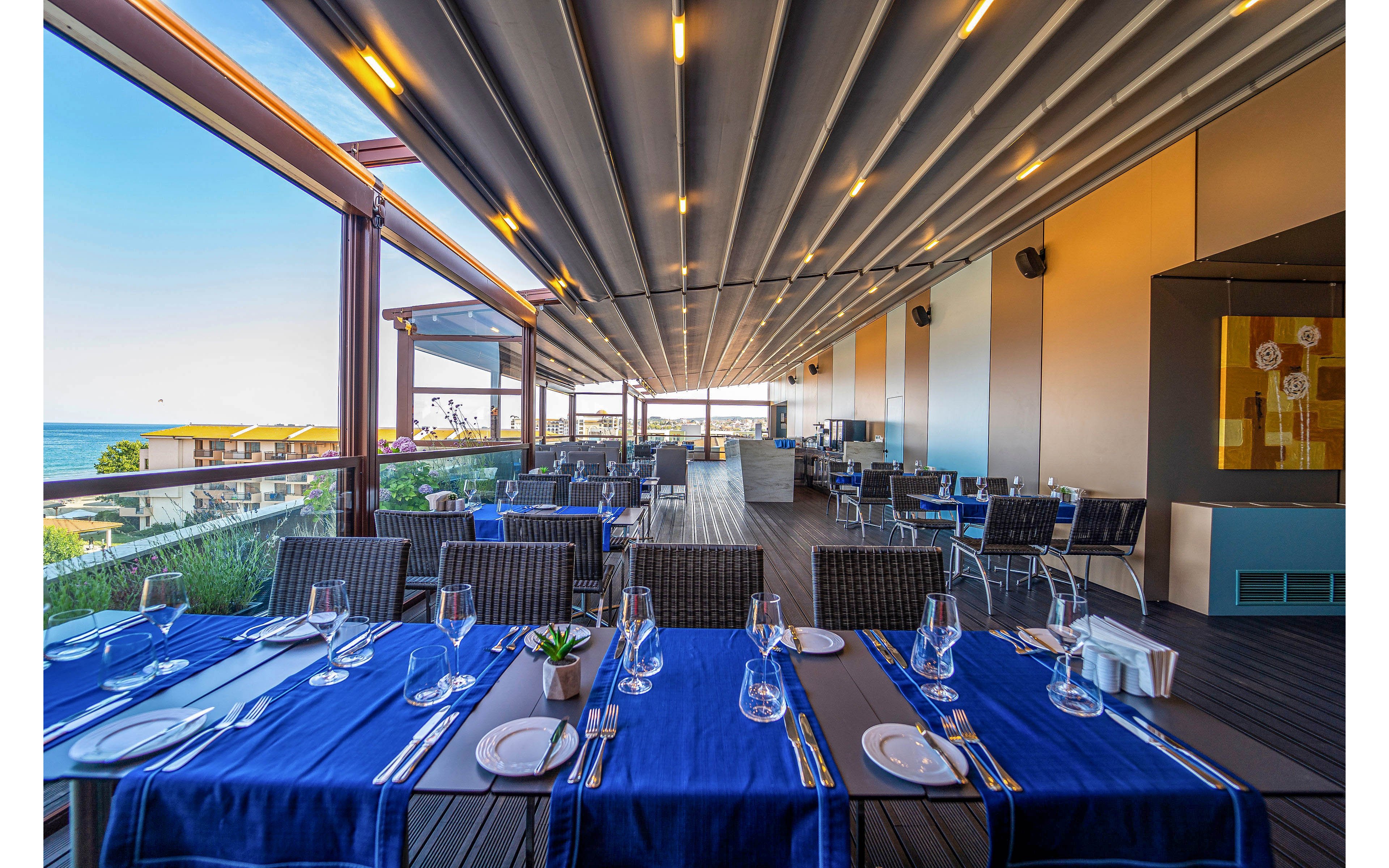 An elegant open-air restaurant terrace is set with neatly arranged tables covered in blue runners and white plates. Rows of wicker-style chairs and glassware create a refined dining atmosphere under a modern retractable roof. Large windows and railings reveal a coastal view with blue sky and sea in the background. The scene suggests a relaxed yet upscale setting prepared for guests at a seaside venue.
