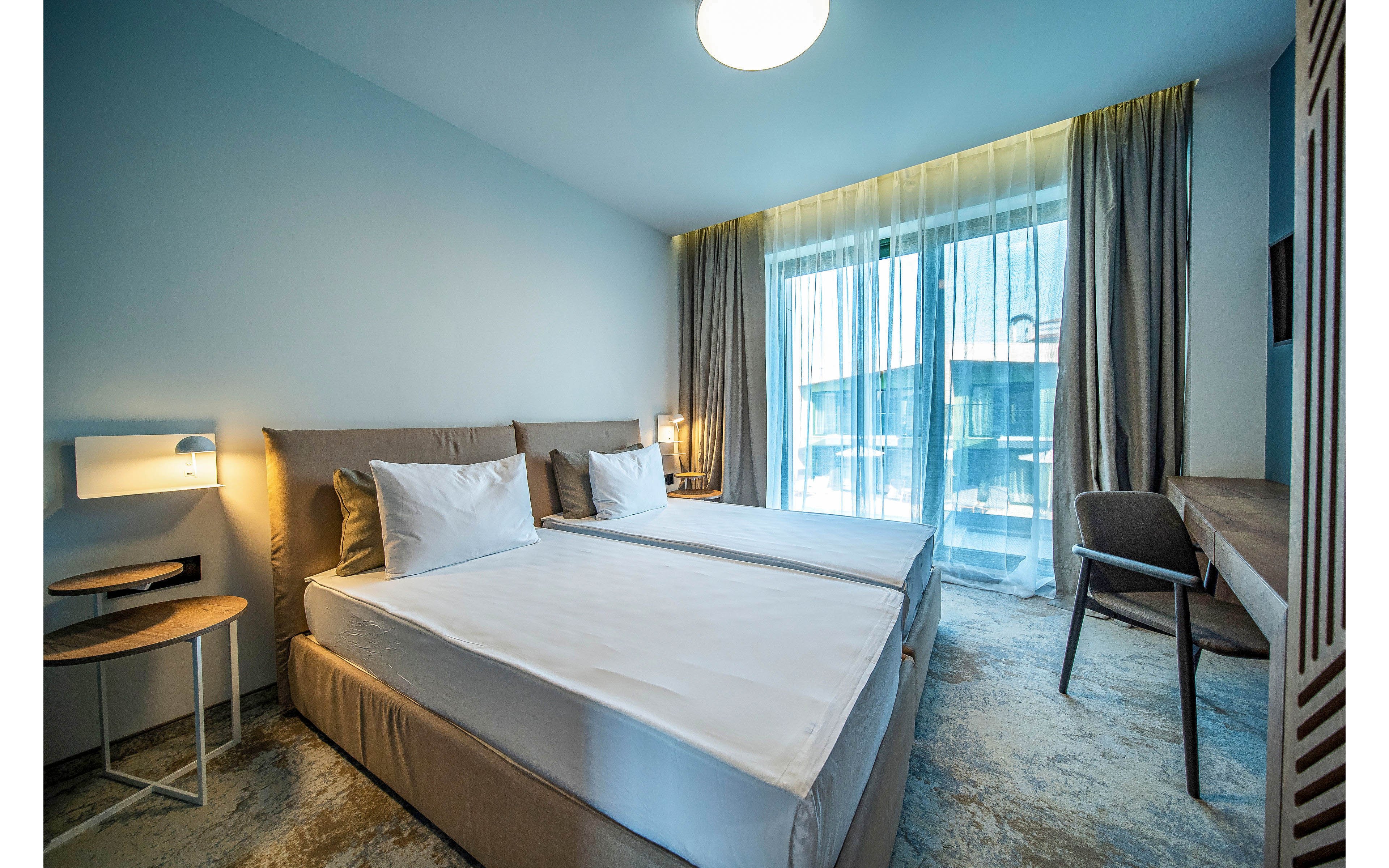 Contemporary hotel room featuring two neatly made twin beds with white linens and neutral upholstered headboards. The space includes bedside tables, a wall-mounted light, and a simple desk with a chair along the right side. Large floor-to-ceiling windows with sheer and blackout curtains let in natural daylight and offer a view of neighboring buildings. Soft blue and beige tones create a calm, minimalist atmosphere suitable for business or leisure travel.