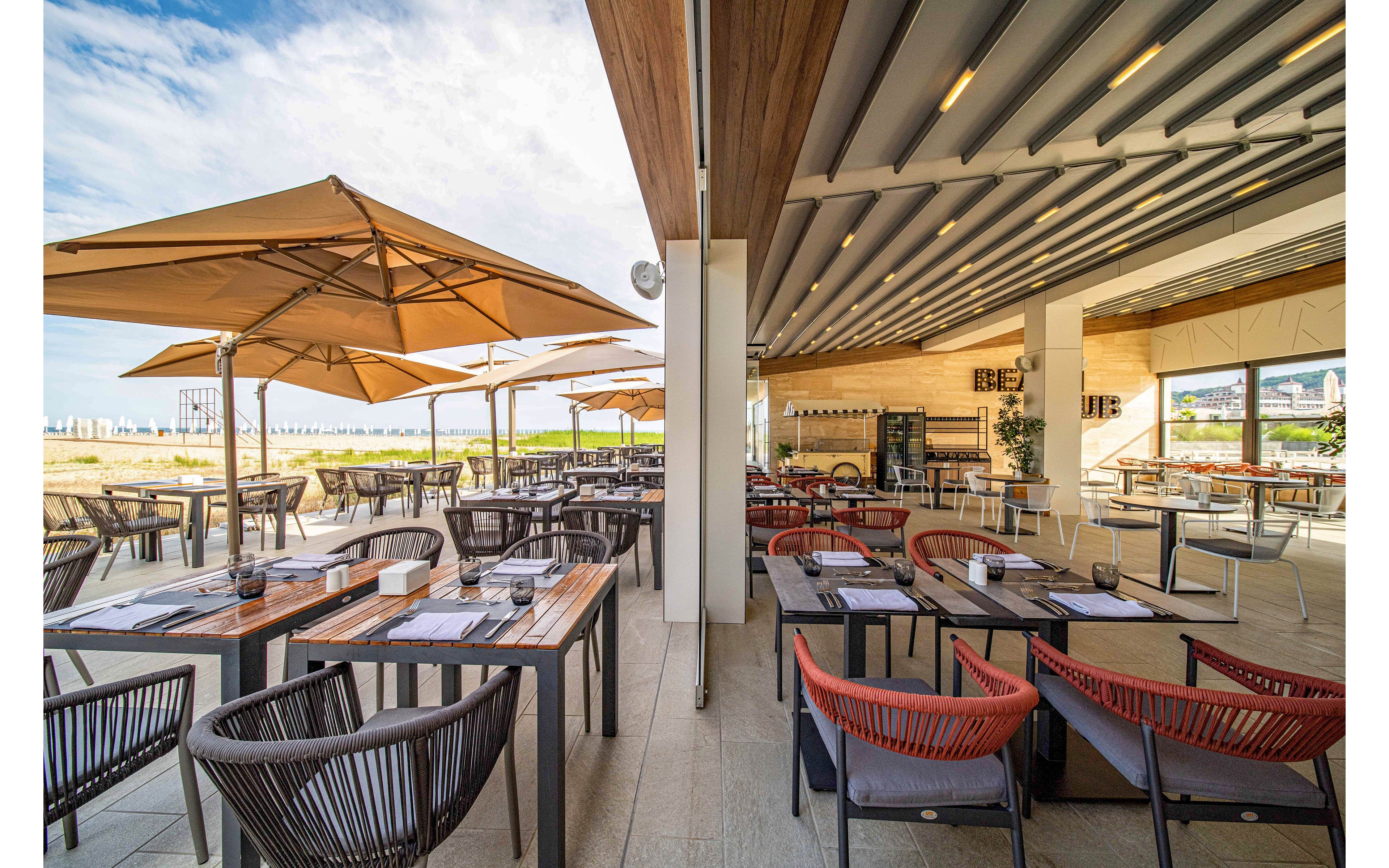 Contemporary outdoor restaurant seating is arranged neatly under a partially covered terrace. Wooden tables with place settings and modern chairs line the space, extending toward large tan umbrellas. The open design looks out onto a bright, sunny landscape with blue sky and distant water or beach. Warm wood tones, neutral flooring, and sleek metal details create a relaxed yet upscale dining atmosphere.