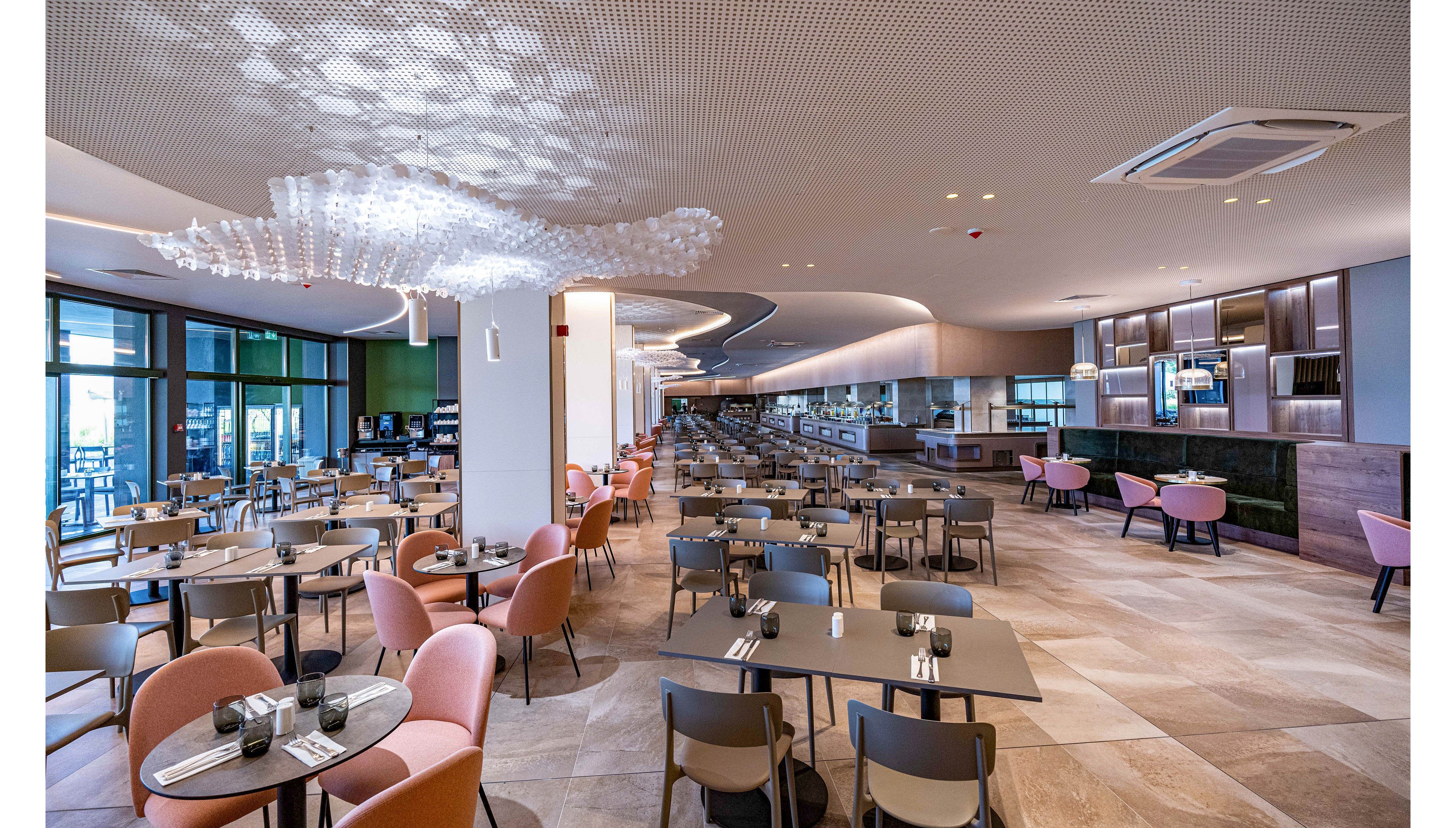 Spacious contemporary restaurant interior with neatly arranged tables and chairs. The dining area features soft pastel seating, tiled floors, and a long open counter along one wall. Large windows on the side bring in natural light, while sculptural ceiling lights add a stylish touch. The space appears clean, organized, and ready to welcome guests.