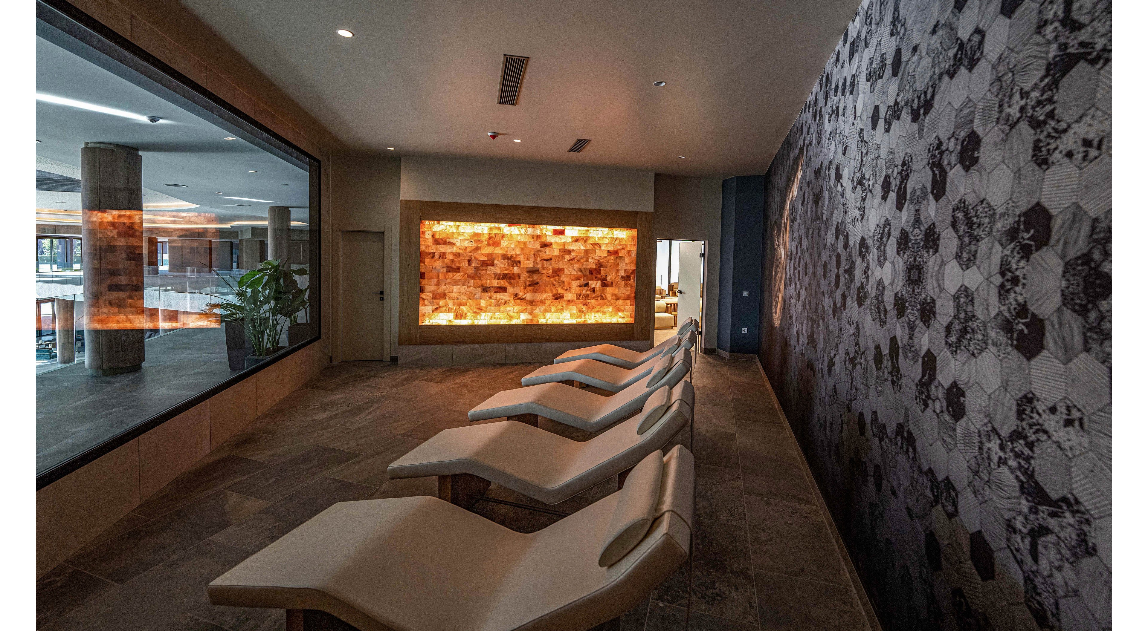 A contemporary spa relaxation room features several reclining lounge chairs arranged in a row. Warm backlit wall panels create a soothing glow against the otherwise dim interior. A large window opens the view to an adjacent area, adding depth and openness to the space. The overall design emphasizes calm, comfort, and modern wellness aesthetics.