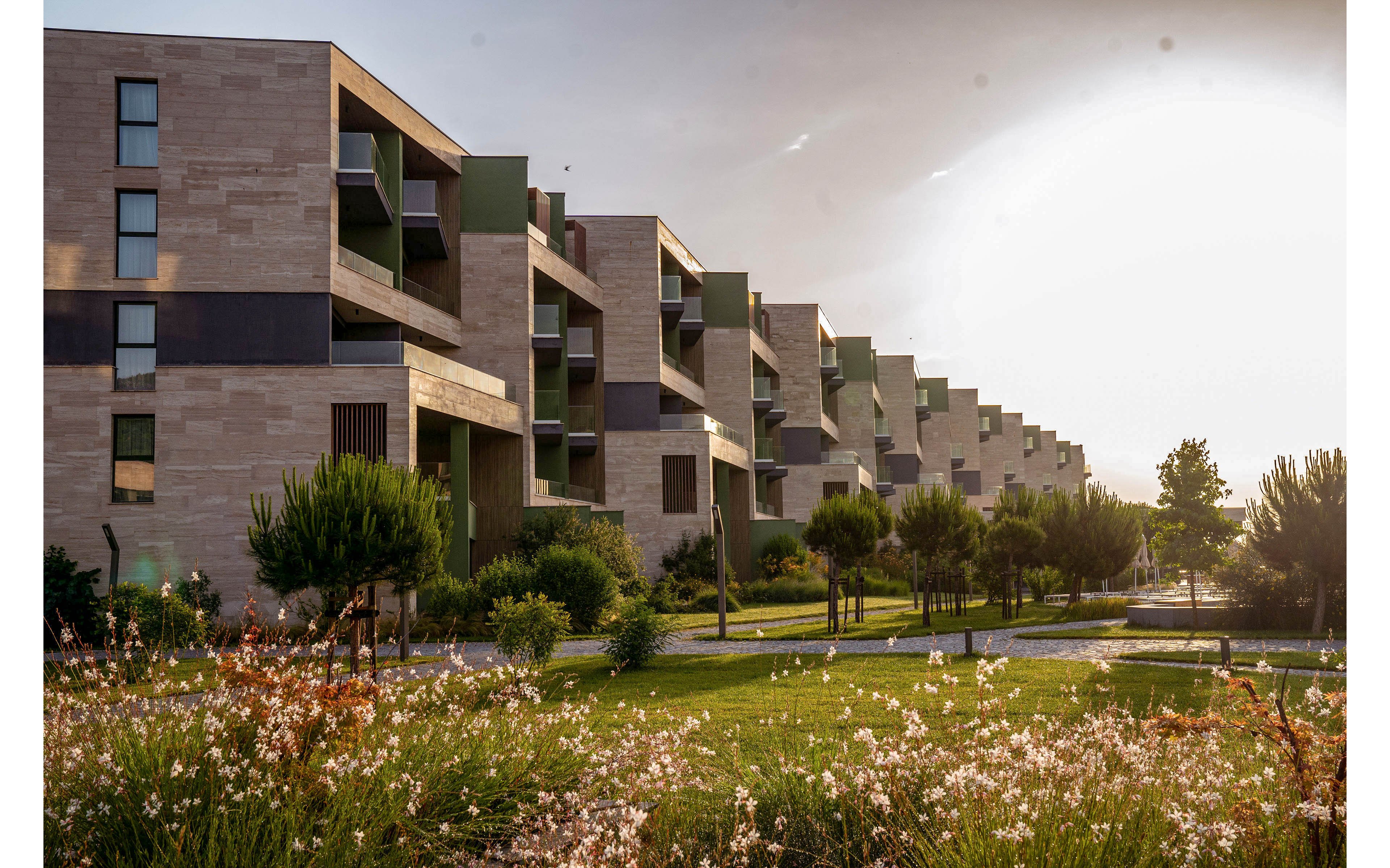 A row of modern apartment buildings stretches into the distance under a warm sunset sky. The architecture features clean lines, balconies, and a mix of stone and green facade panels. In the foreground, a landscaped garden with flowering plants, shrubs, and small trees softens the urban setting. The overall scene conveys a peaceful residential neighborhood with contemporary design and ample green space.