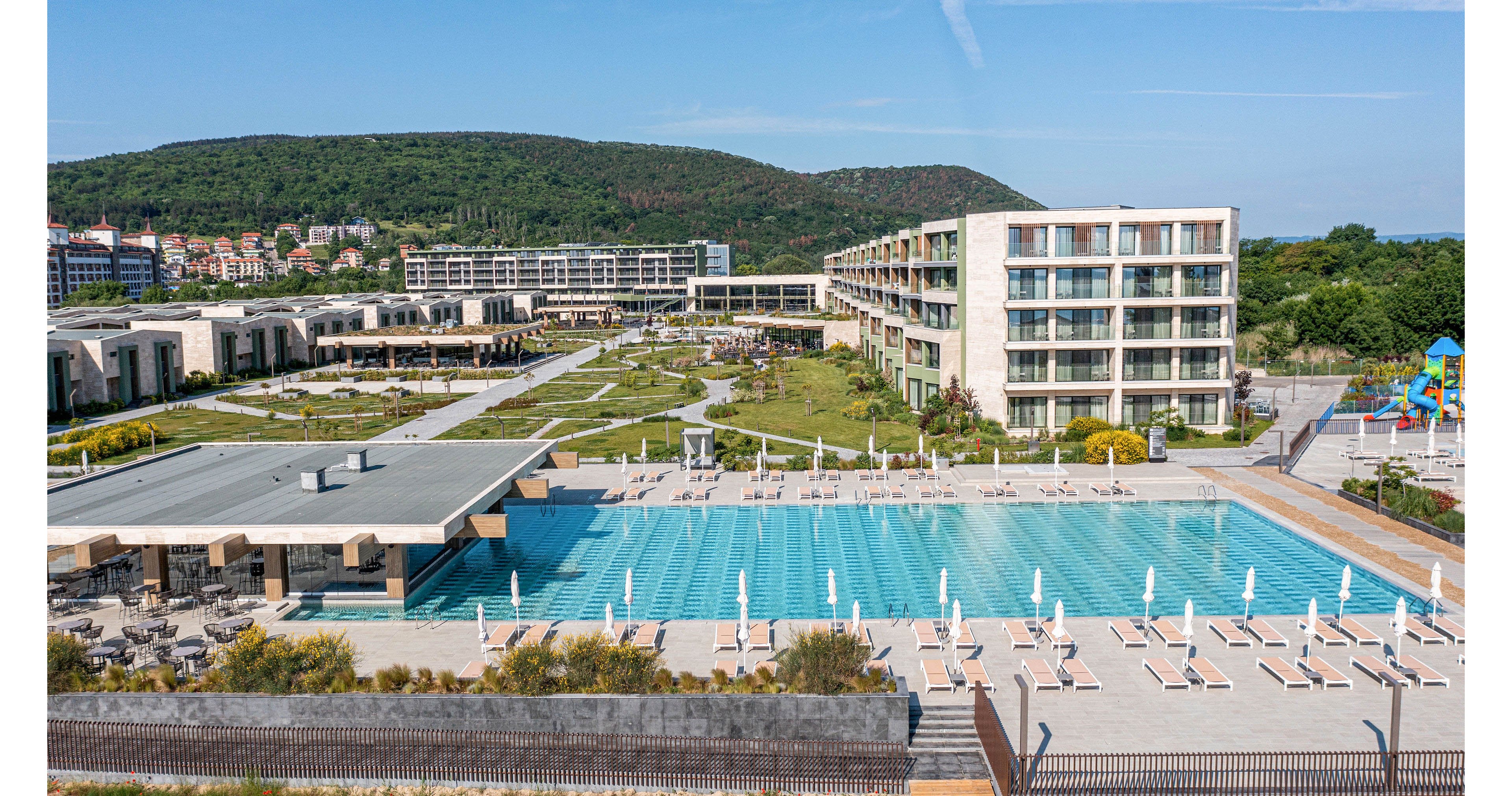 A contemporary resort complex features a large rectangular outdoor swimming pool lined with white sun loungers and umbrellas. Several multi-story hotel buildings and low-rise villas surround the pool, set within landscaped green grounds. In the background, forested hills and a small town are visible under a clear blue sky. The scene suggests a sunny, relaxing vacation destination with ample outdoor amenities.