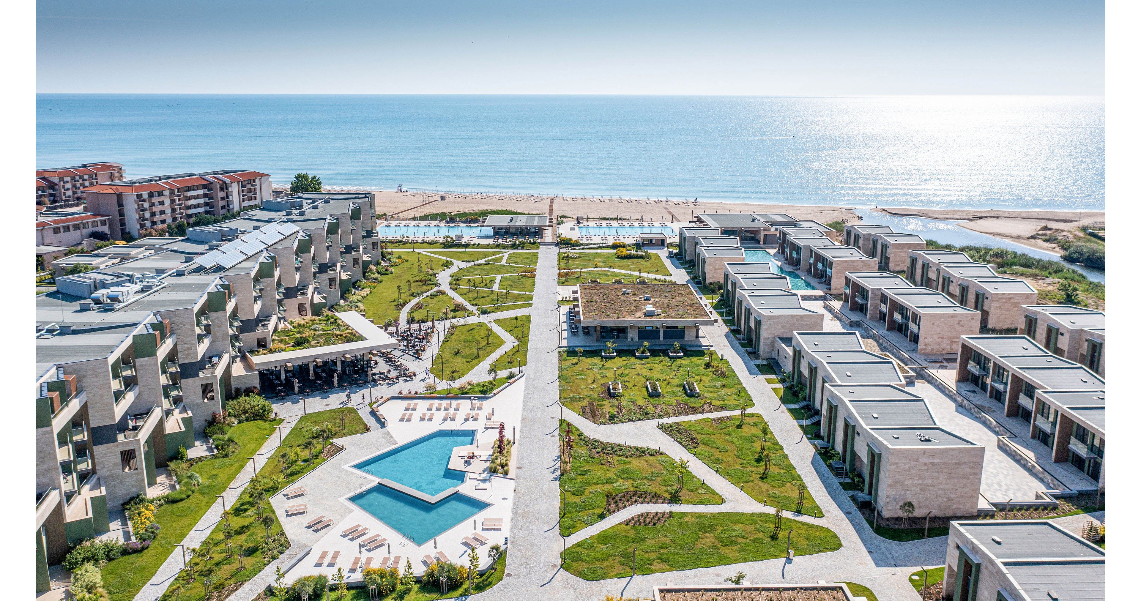 A contemporary seaside resort stretches along a sandy coastline, featuring multiple pools and landscaped green spaces. Modern multi-story buildings line the left side, while rows of private villa-style units occupy the right. Pathways, lawns, and sun loungers create an inviting leisure environment under clear blue skies. The calm sea in the background emphasizes the coastal vacation setting.