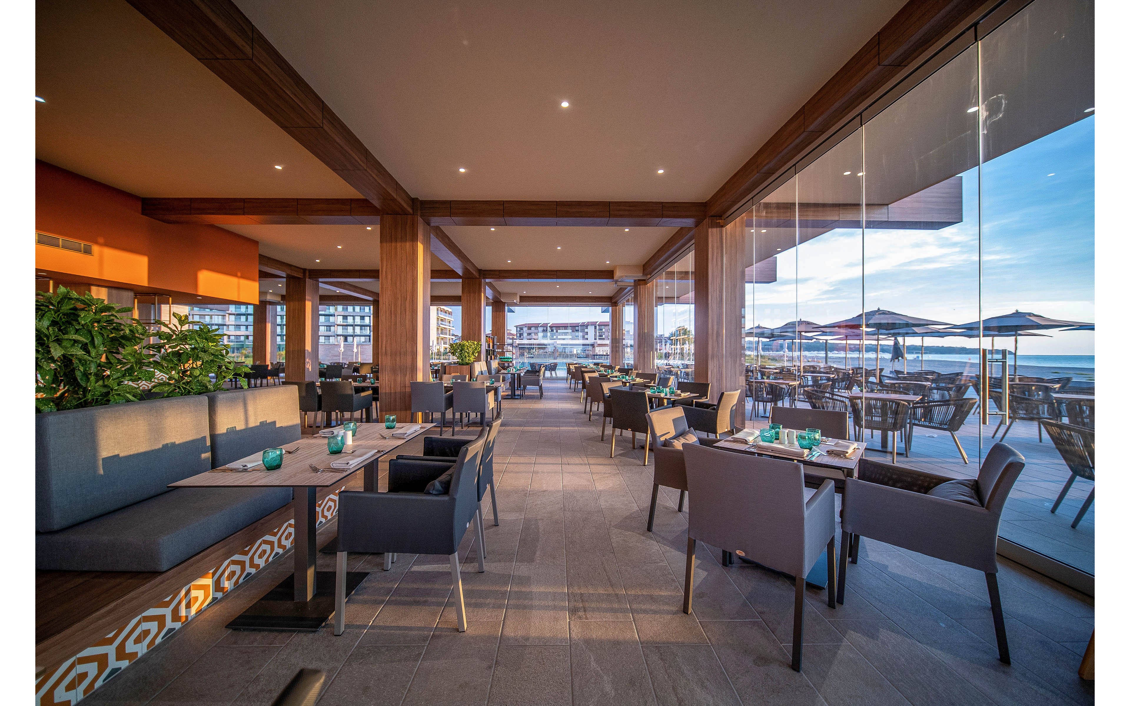 A contemporary open-air restaurant features neatly arranged tables and cushioned seating under a wooden ceiling. Large floor-to-ceiling glass windows reveal an outdoor terrace with umbrellas and a view of the water. Soft evening light creates long shadows and a warm, inviting atmosphere. The space appears calm and ready for guests, emphasizing comfort, style, and scenic oceanfront dining.