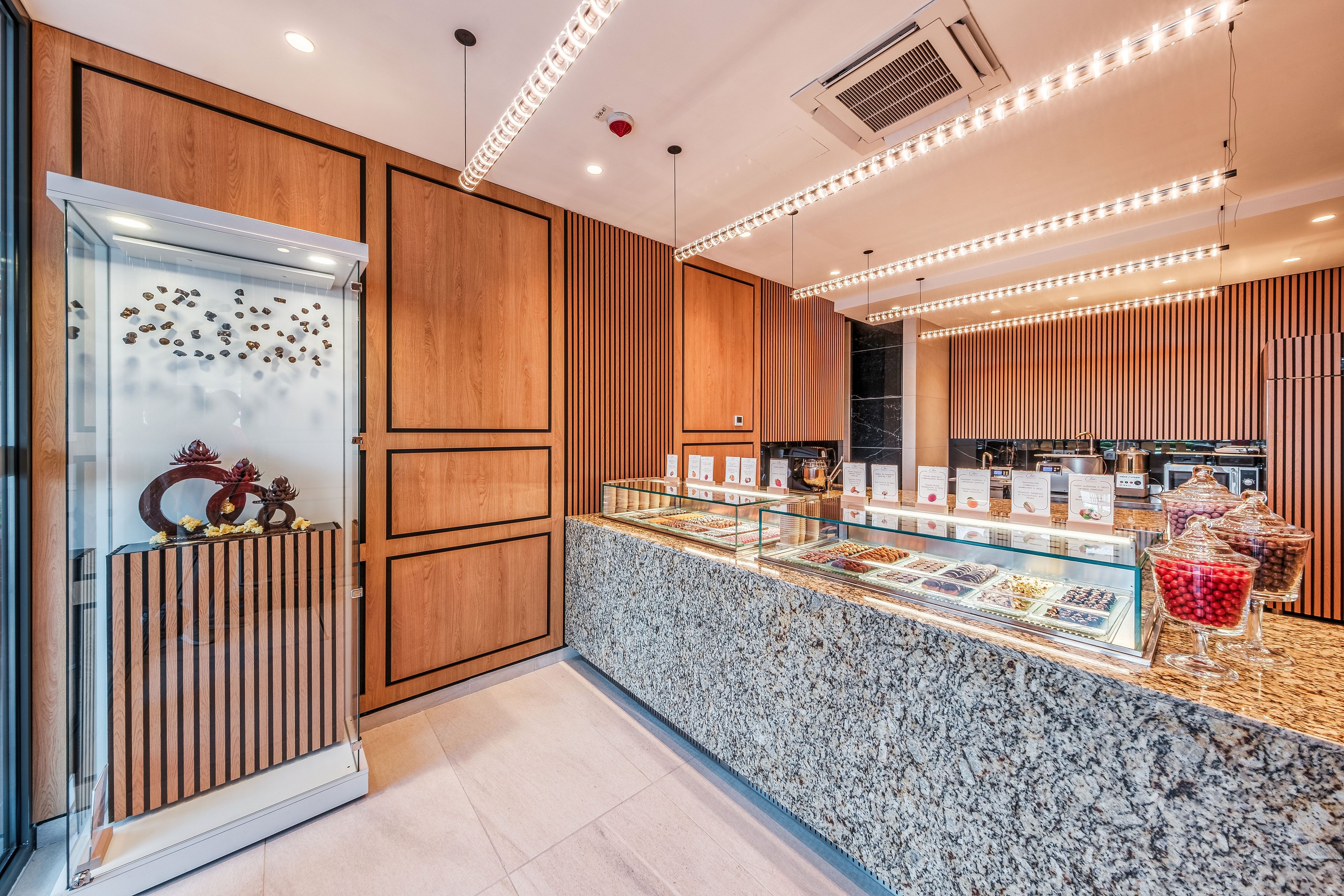 Bright, contemporary confectionery shop interior featuring a long granite display counter filled with assorted chocolates and sweets. Wooden wall panels and vertical slats create a warm, upscale atmosphere, complemented by linear ceiling lights. A glass showcase on the left displays decorative chocolate sculptures, adding an artistic touch. Clear signage and product cards line the counter, suggesting a curated selection of premium treats.