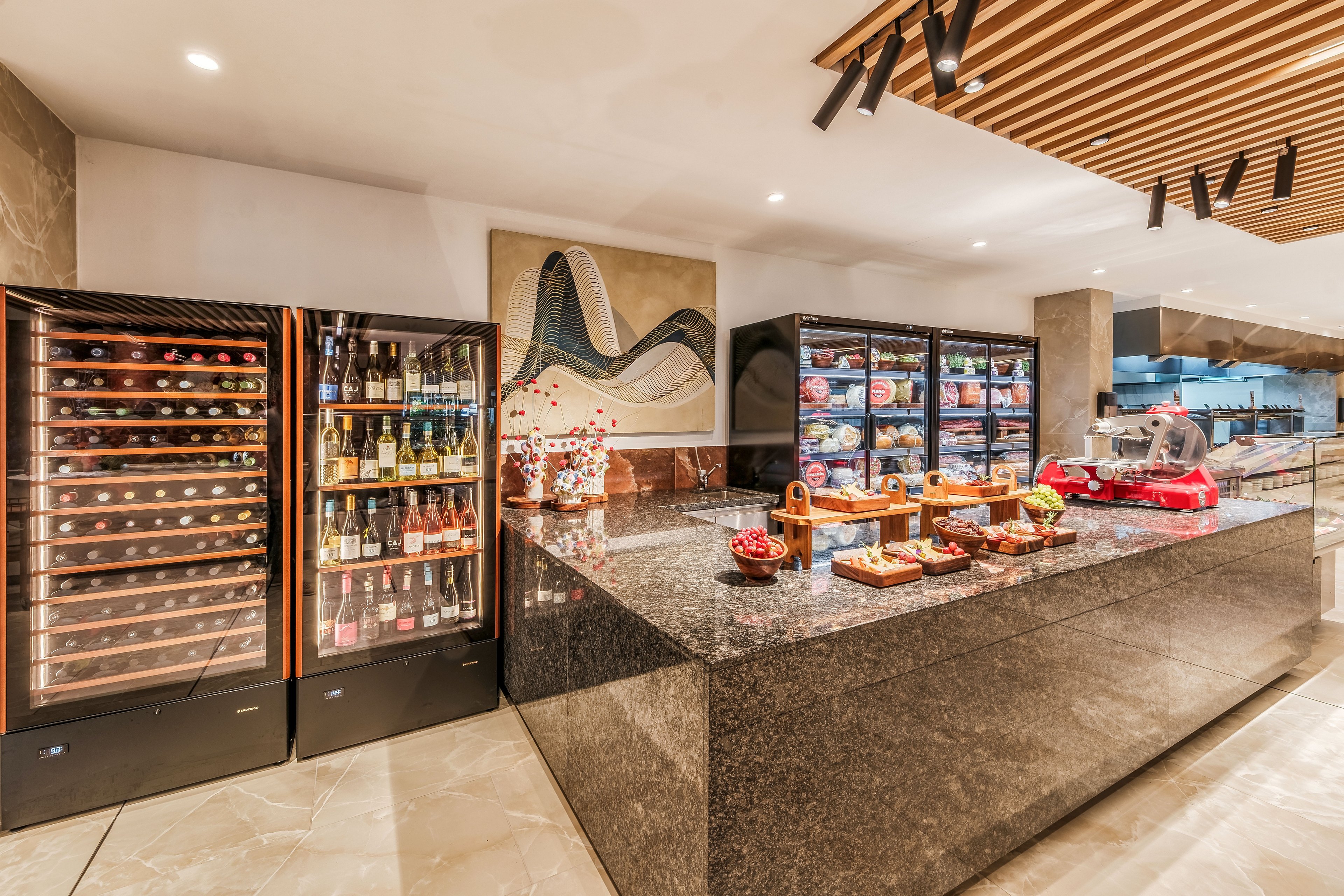A sleek granite buffet counter is set in a contemporary restaurant or hotel dining area. Refrigerated display cases and wine fridges showcase bottles, beverages, and assorted chilled foods. The countertop features neatly arranged pastries, bread, fruit, and other appetizers, with a meat slicer visible to the right. Warm lighting, wood ceiling slats, and abstract wall art create an upscale, inviting atmosphere.