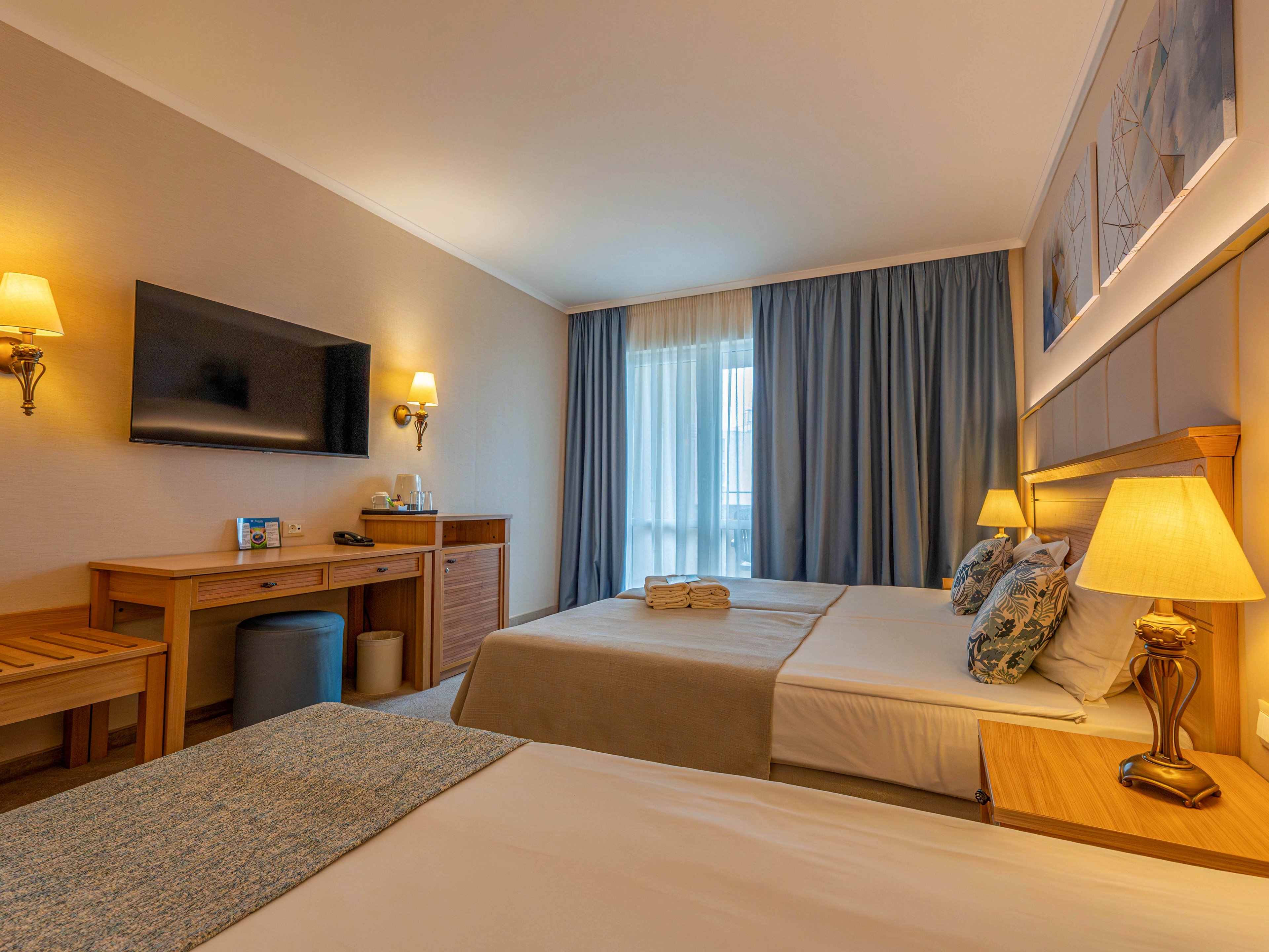 A warm, inviting hotel room features two neatly made beds with soft lighting from bedside lamps. A large flat-screen television is mounted on the wall above a wooden desk and stool, with amenities arranged on the surface. Floor-to-ceiling curtains cover a window that lets in natural daylight, complementing the neutral tones and blue accents in the decor. The overall atmosphere is calm, comfortable, and suitable for relaxation or a short stay.