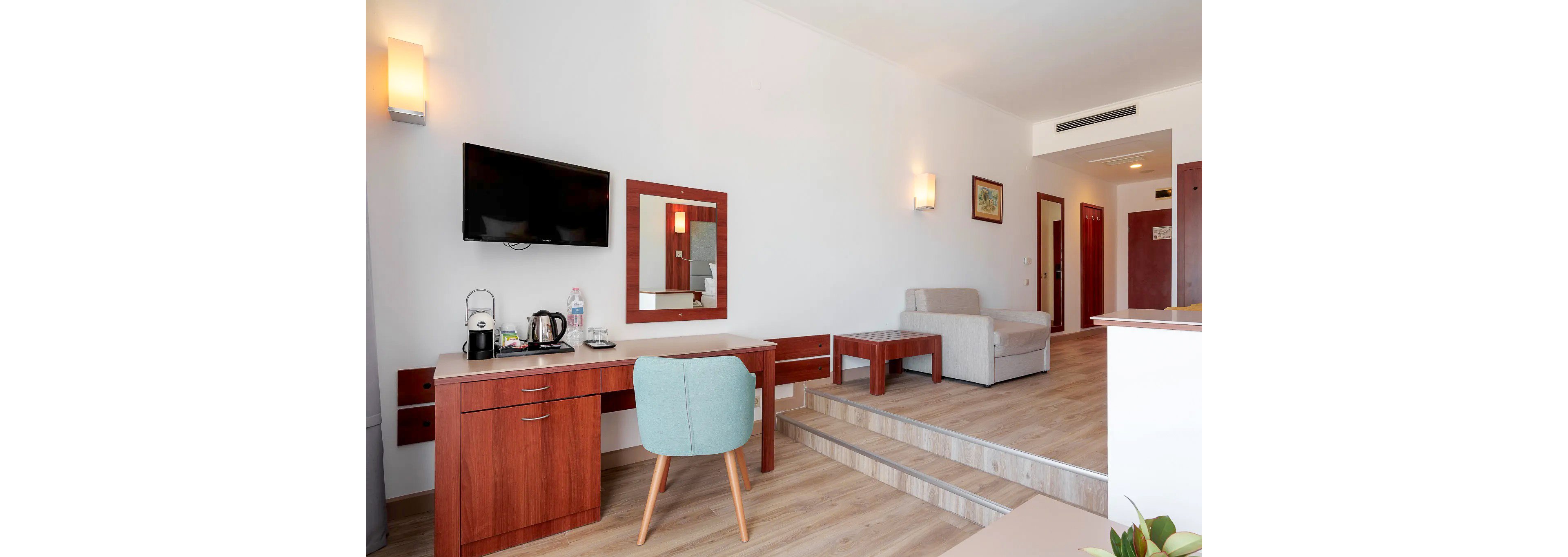 A bright and modern hotel room featuring a wooden desk with a teal chair, a mounted flat-screen TV, and a cozy seating area with a sofa. The room has a clean and minimalistic design with light-colored walls and wooden flooring. A kettle, water bottle, and coffee cups are placed on the desk, adding a touch of convenience. The space is well-lit with wall-mounted lights and natural light from an unseen window.