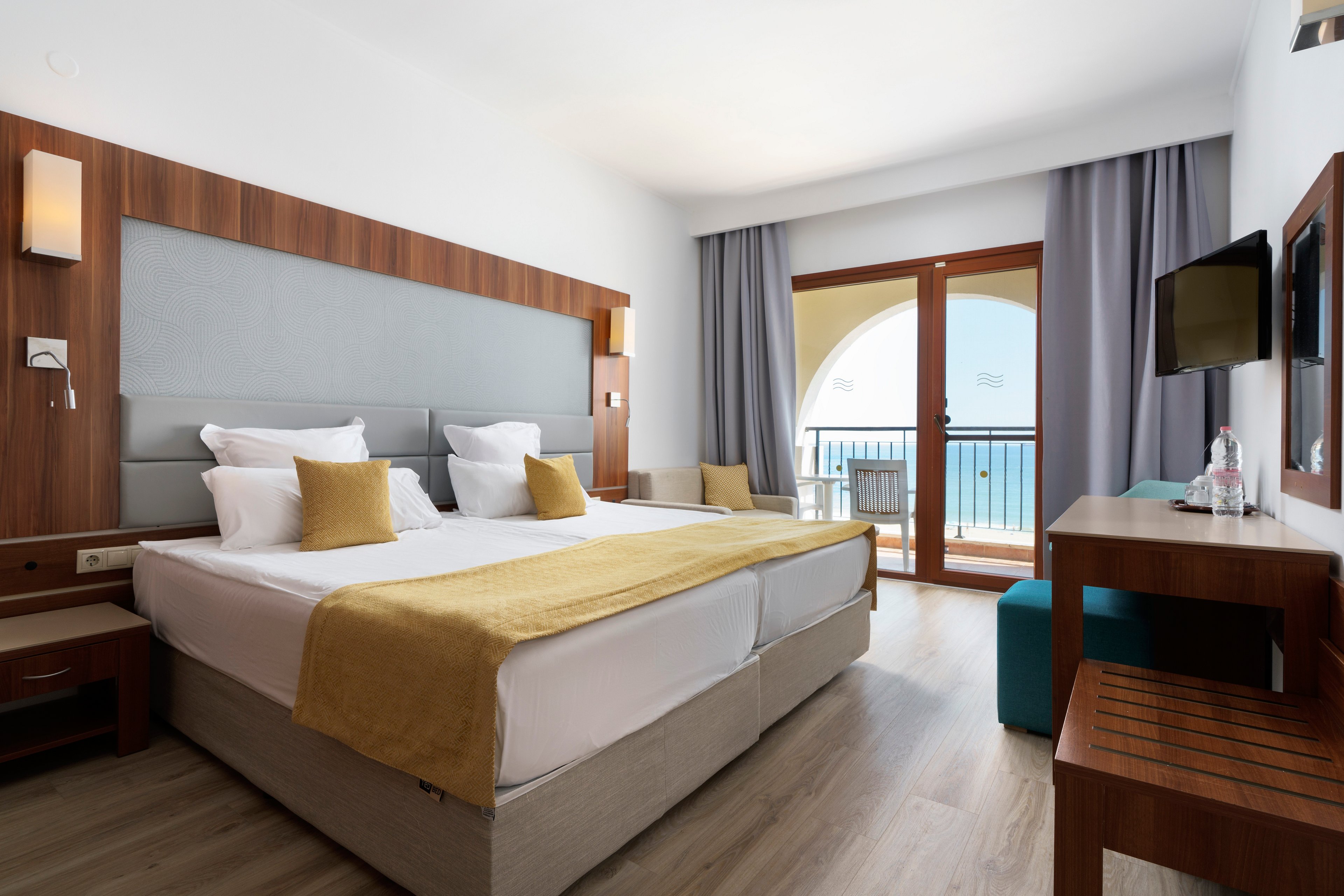 A contemporary hotel bedroom features a large double bed with white linens and mustard yellow throws and cushions. The room includes wooden furniture, a wall-mounted television, and a small desk with bottled water and glasses. Floor-to-ceiling glass doors open onto a balcony with a view of the sea framed by an arched opening. Soft gray curtains and light wood flooring create a clean, minimalist atmosphere.