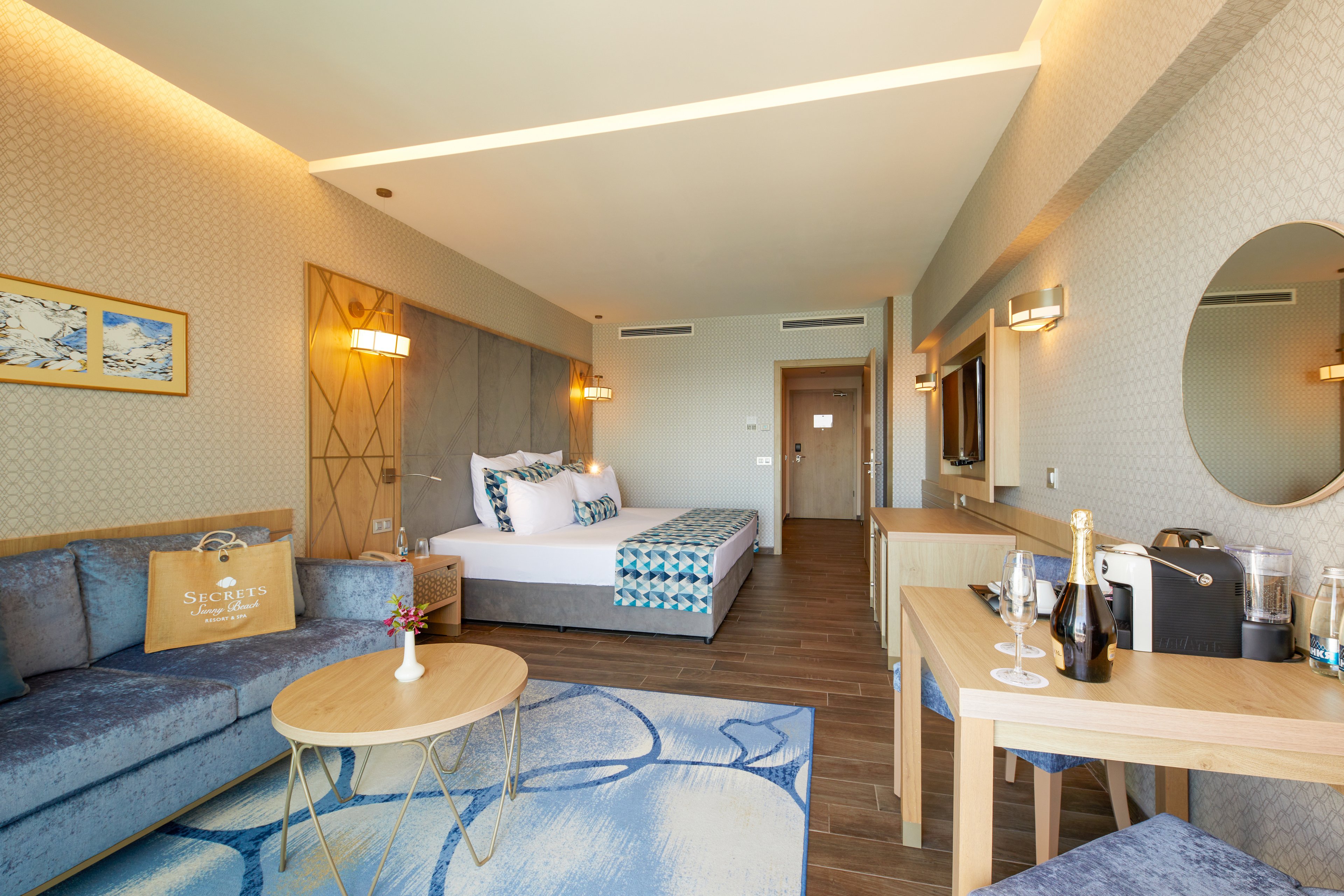 A spacious and modern hotel room featuring a comfortable bed with patterned bedding, a seating area with a blue sofa, and a wooden table. The room is well-lit with warm lighting and decorated with abstract wall art. A bottle of champagne and glasses are placed on the dining table, adding a touch of luxury. The room has a serene and inviting atmosphere, with a mix of neutral and blue tones.
