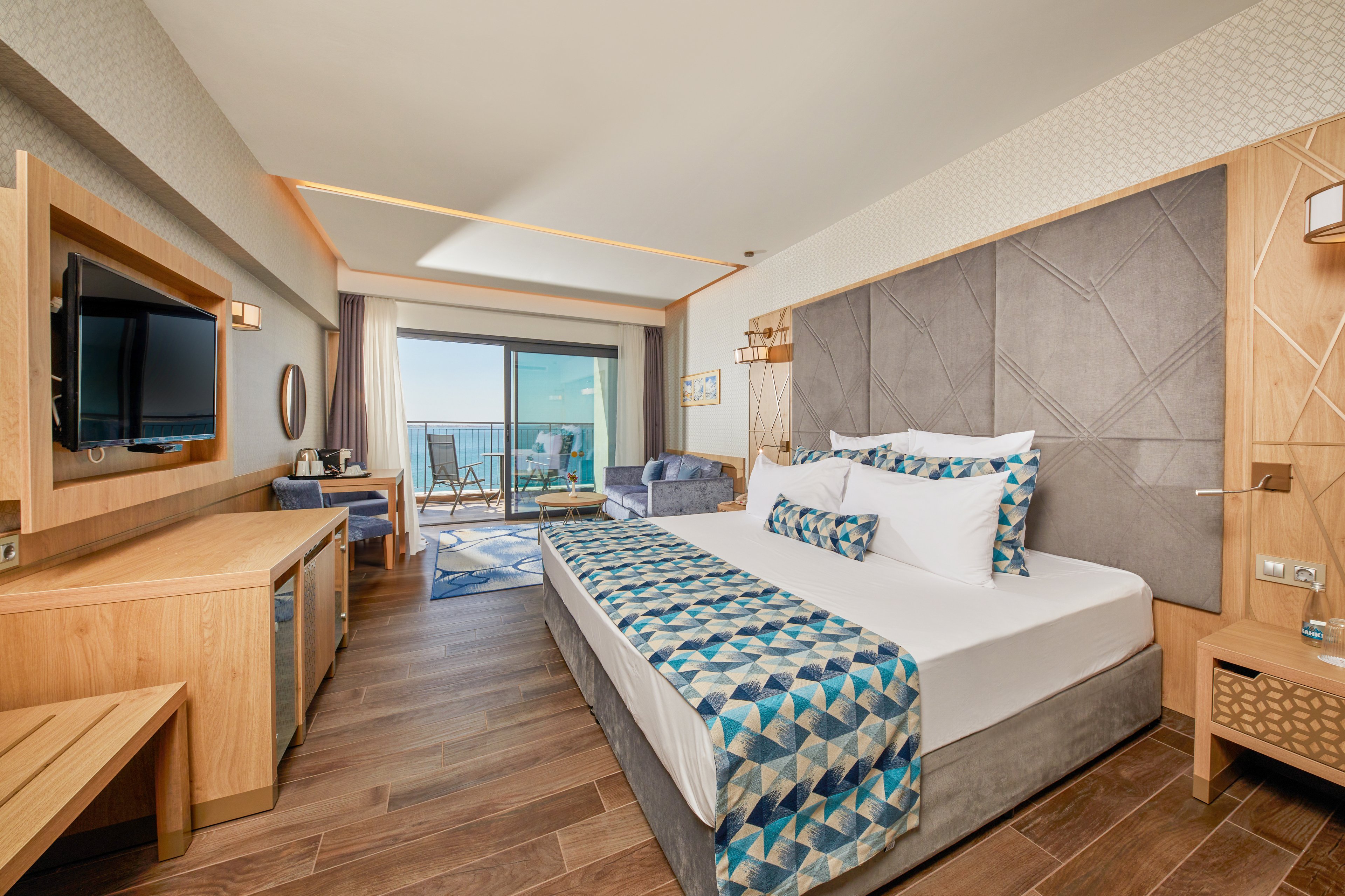 A contemporary hotel room featuring a neatly made bed with geometric-patterned bedding, wooden furniture, and a flat-screen TV. The room opens to a balcony with a scenic ocean view, complemented by natural light streaming through large glass doors. The setting is cozy and inviting, ideal for relaxation or vacation stays.