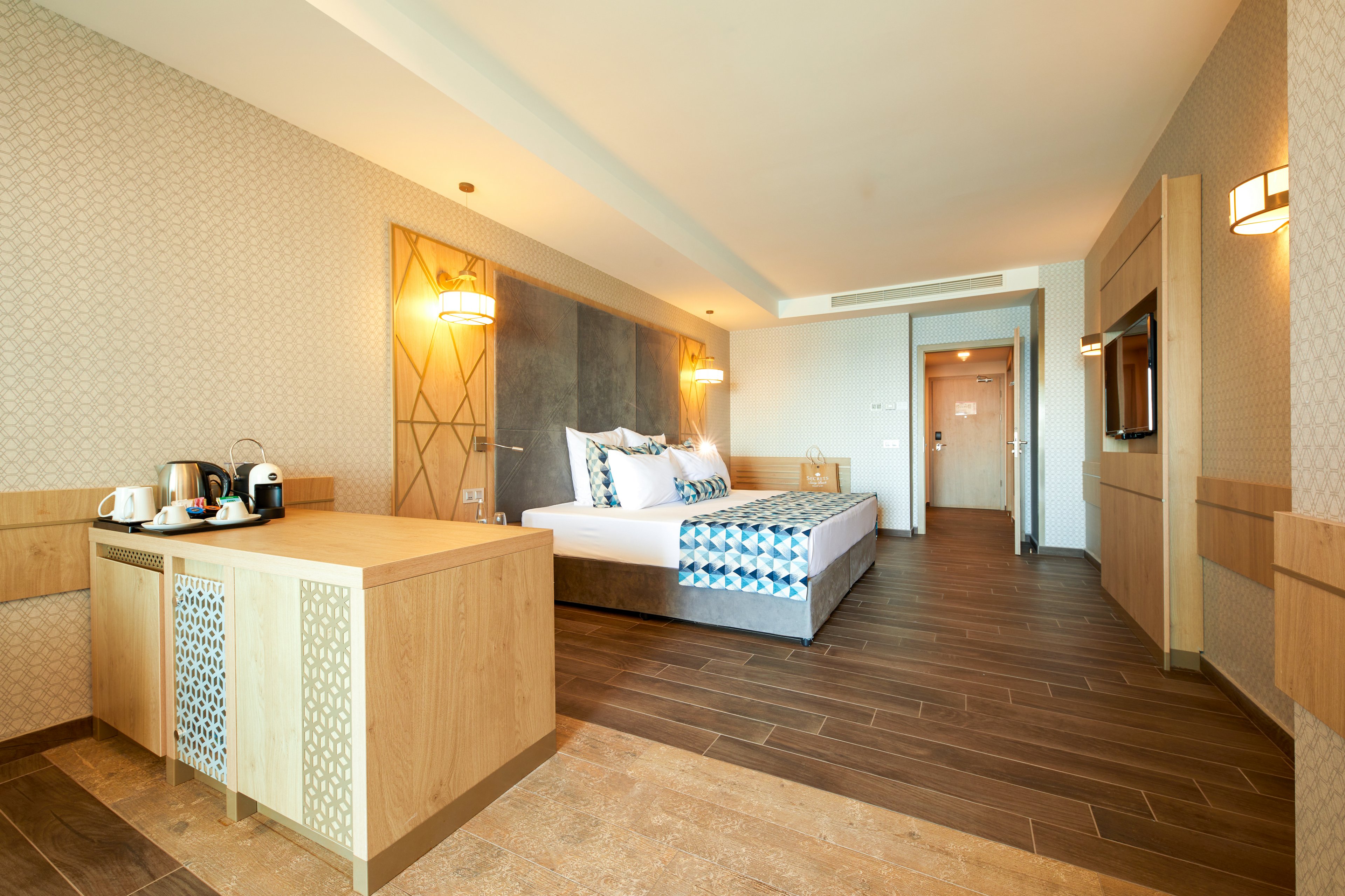 A spacious and modern hotel room featuring a neatly made bed with patterned bedding, warm lighting, and wooden flooring. The room includes a small kitchenette area with a coffee machine and cups. The neutral color palette and soft lighting create a welcoming and relaxing atmosphere.