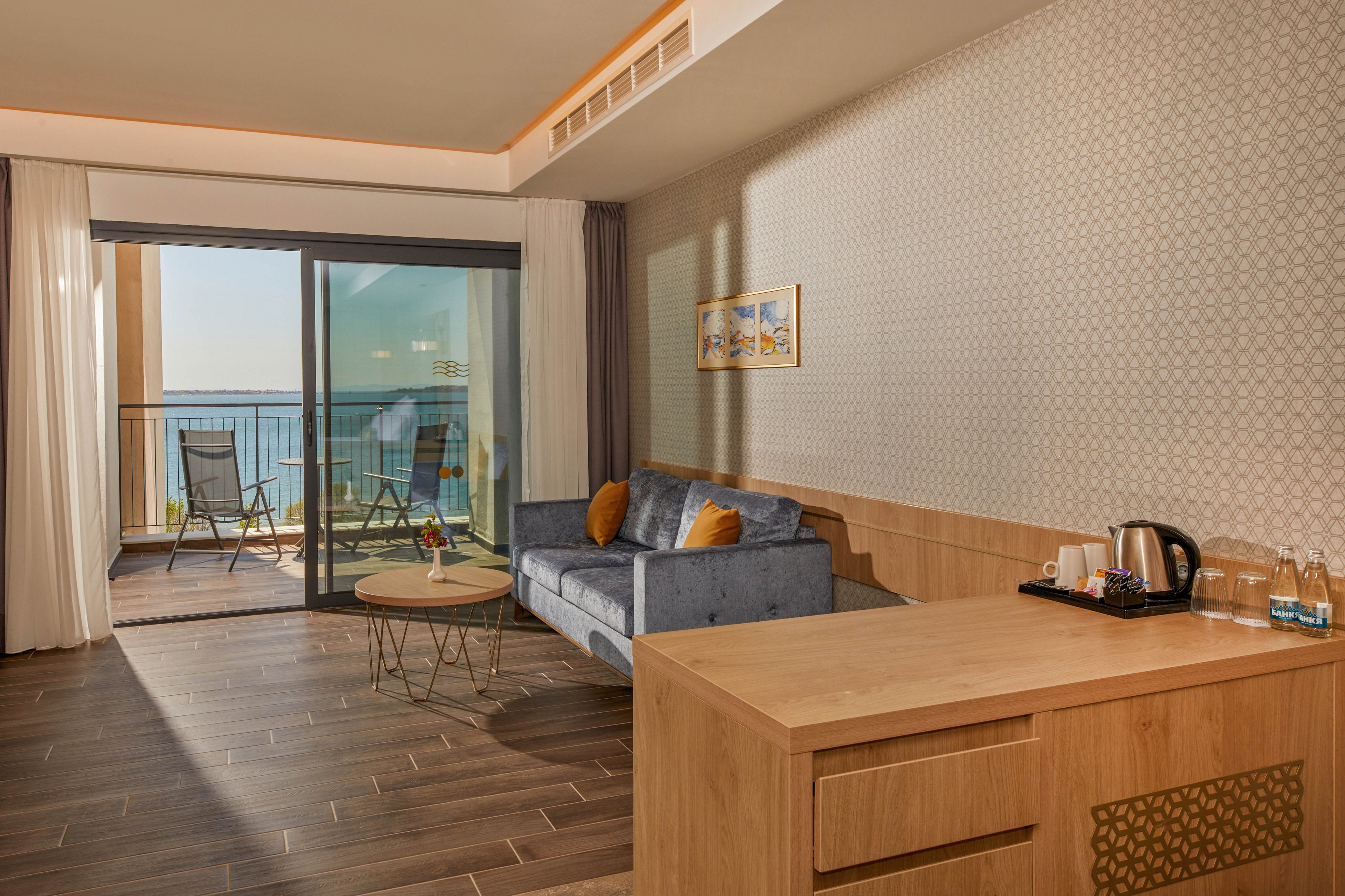 A contemporary hotel suite features a cozy seating area with a blue sofa and accent pillows. Large sliding glass doors open onto a balcony with two chairs and a small table overlooking the sea. Warm wooden furniture, including a desk with a kettle and glasses, contrasts with the patterned wall and tiled floor. Soft natural light and neutral tones create a calm, inviting atmosphere ideal for relaxation.